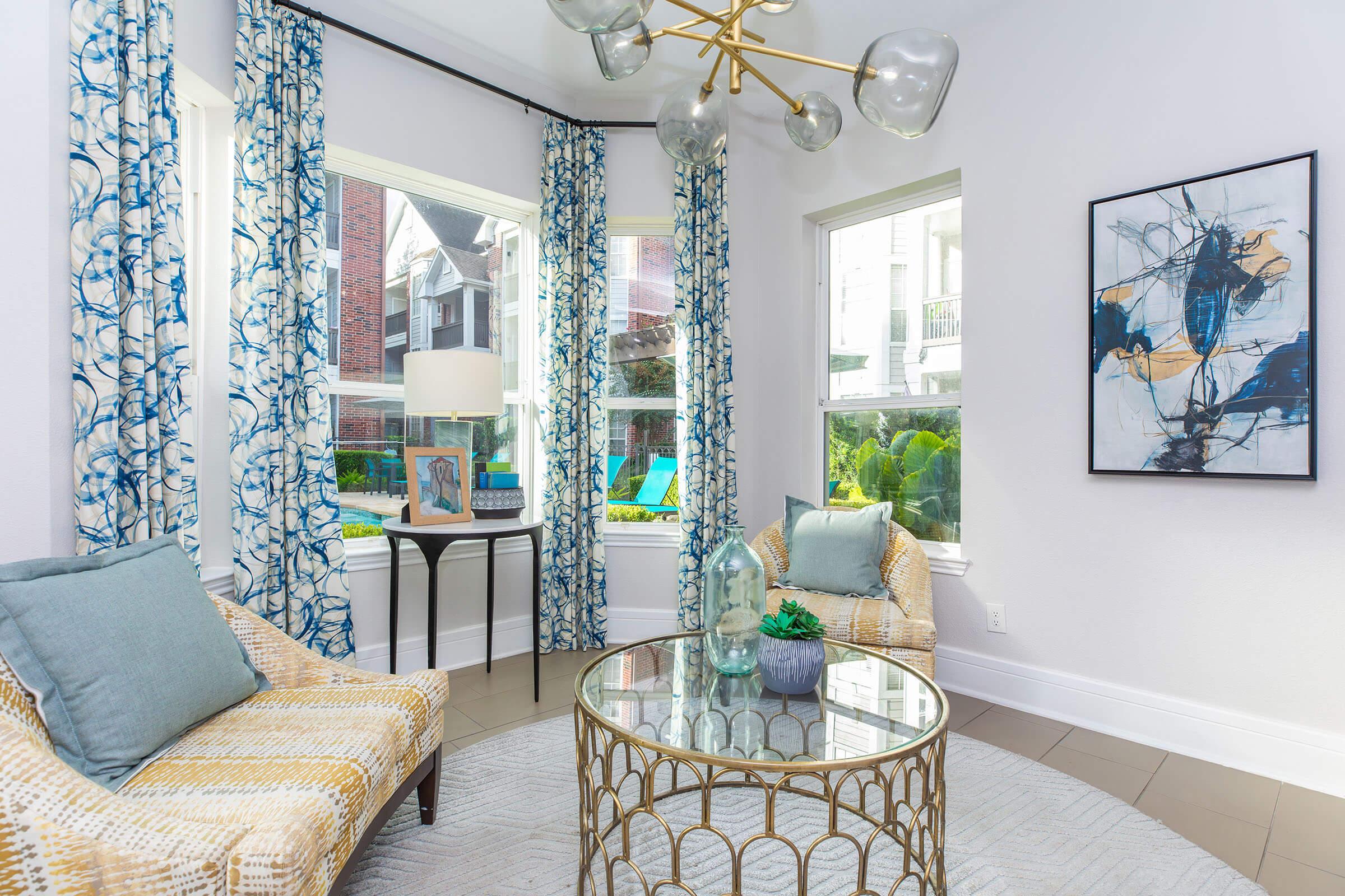 A bright and modern living space featuring two patterned chairs, a glass-top coffee table with decorative items, and large windows adorned with blue and white curtains. A stylish light fixture hangs from the ceiling, and a framed abstract painting adds artistic appeal to the room.