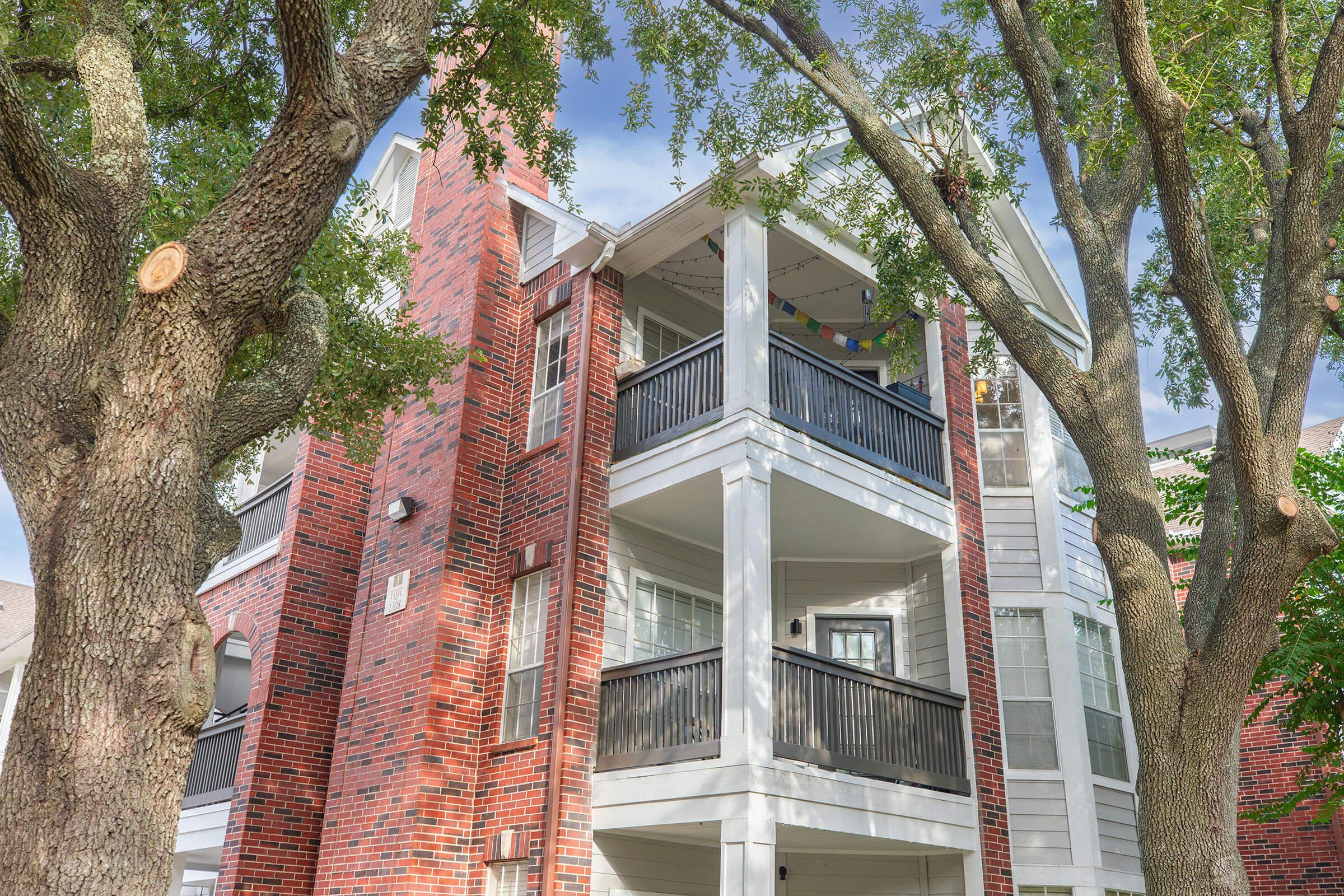 A multi-story red brick apartment building with white accents, featuring balconies on the second and third floors. Surrounding trees provide shade, with green leaves visible against a clear blue sky. Decorative lights hang from the balcony, adding a cheerful touch to the exterior.