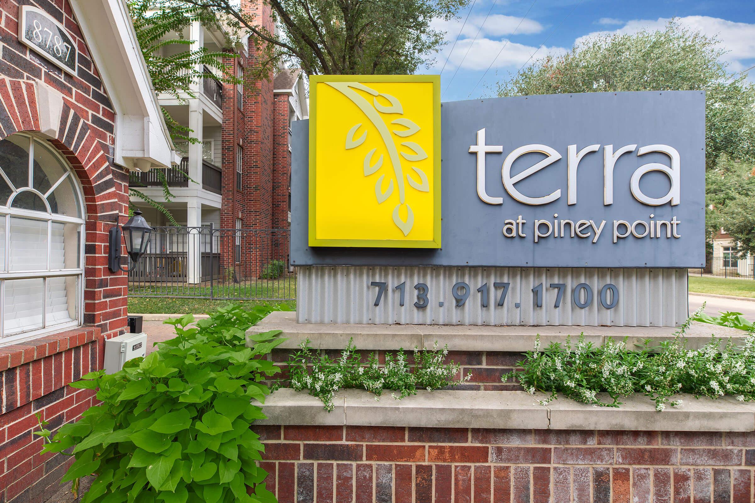 Signage for "terra at piney point" with a logo featuring a leaf design. The sign includes contact information: phone number 713-917-1700. The background features a brick wall with greenery and part of a residential building visible.