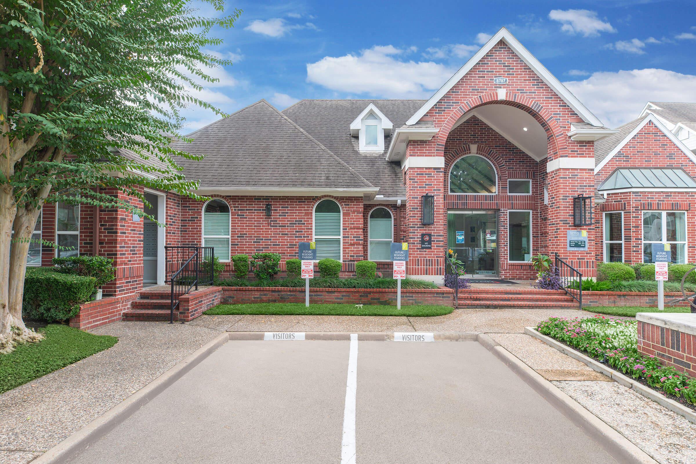 A brick building with a large arched entrance, surrounded by landscaped greenery and parking spaces. The structure features large windows and a gabled roof, set against a clear blue sky. There are signs for designated parking spots in front of the building.