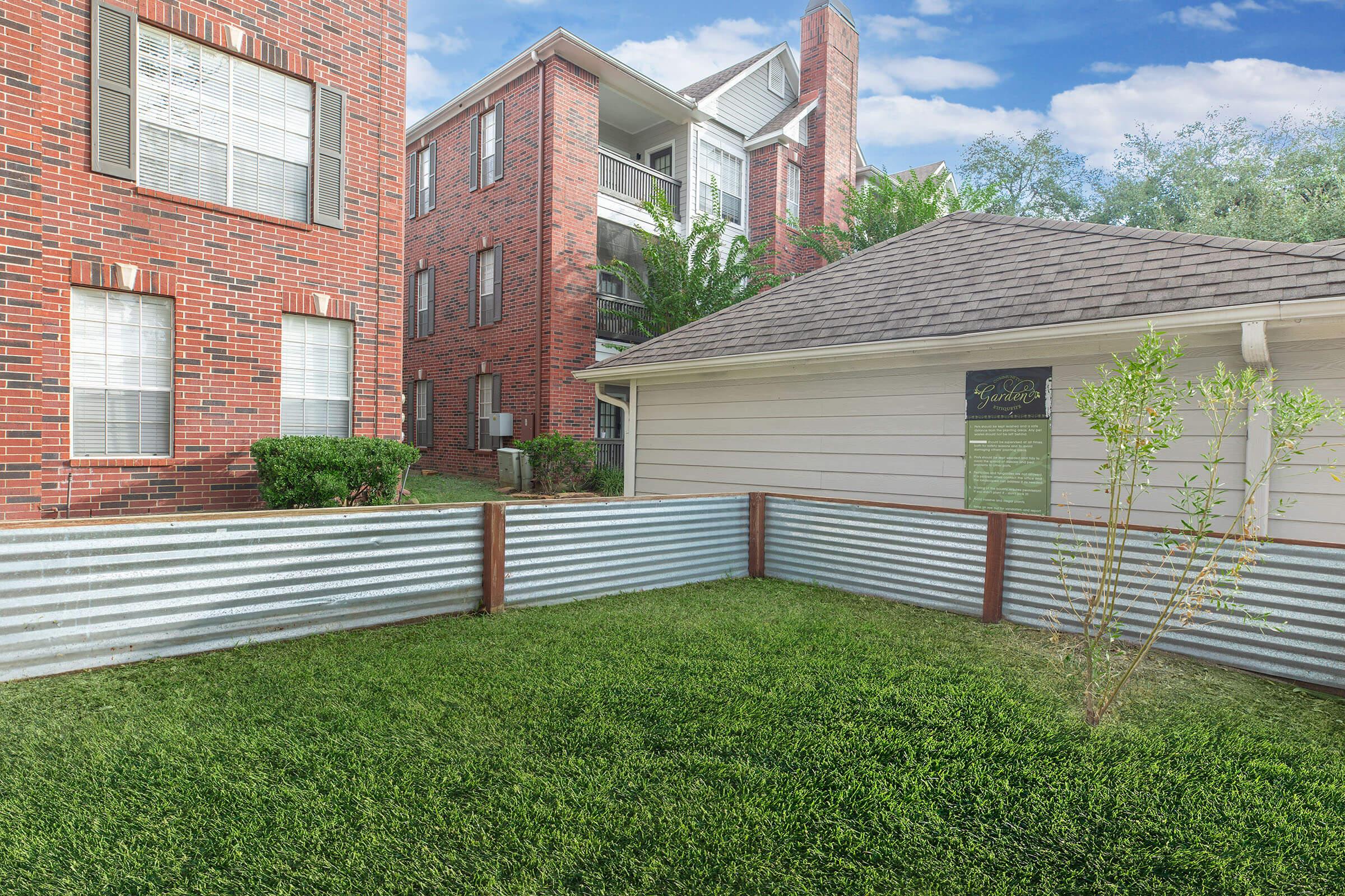 A landscaped area featuring green grass bordered by corrugated metal fencing, surrounded by brick buildings and a white structure with a sign. The sky is clear with a few clouds, creating a peaceful outdoor setting.