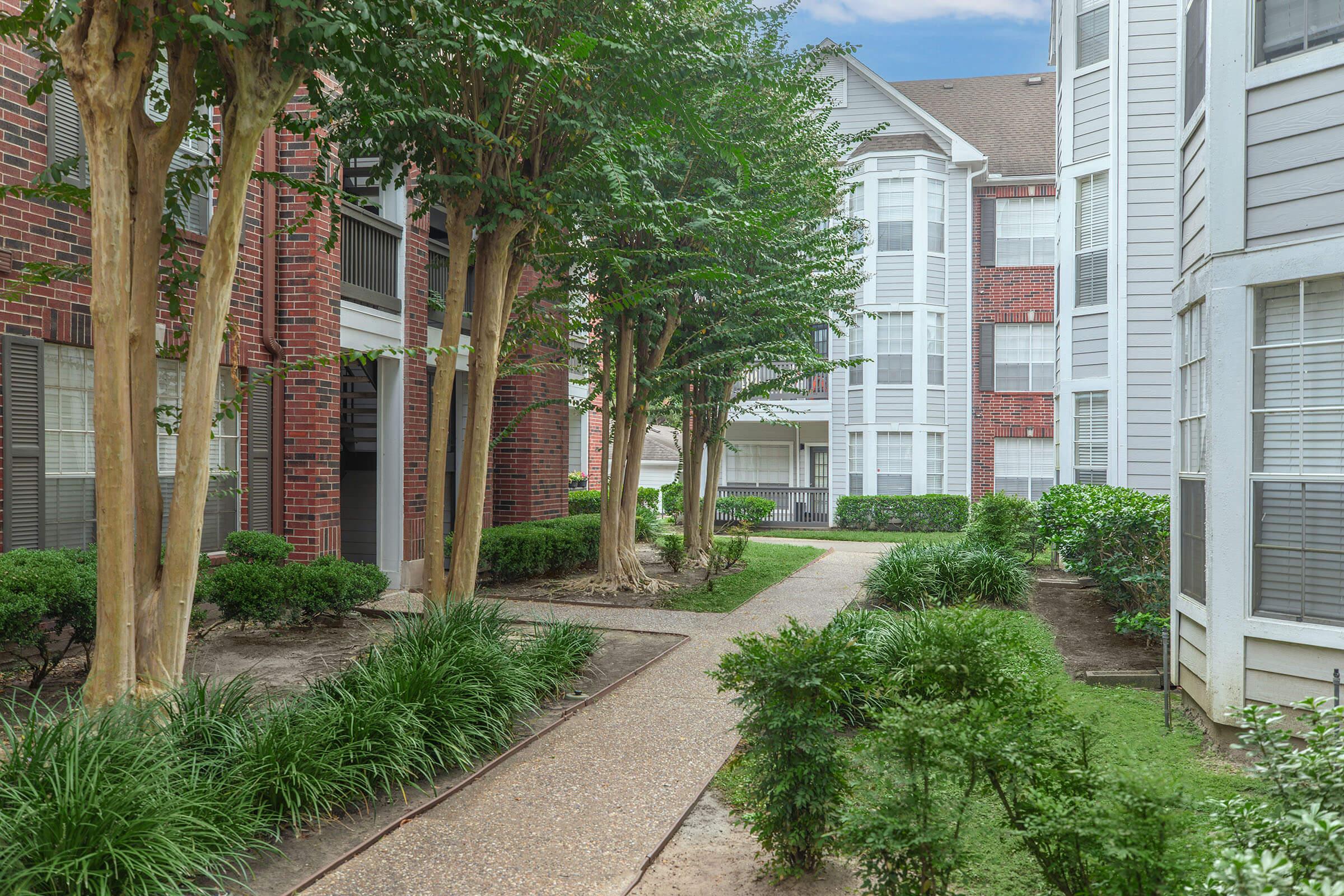 A landscaped apartment courtyard featuring a gravel path, surrounded by neatly trimmed hedges and small trees. The buildings on either side are a mix of brick and light-colored siding, with large windows allowing natural light into the spaces. A peaceful and inviting outdoor ambiance is conveyed.