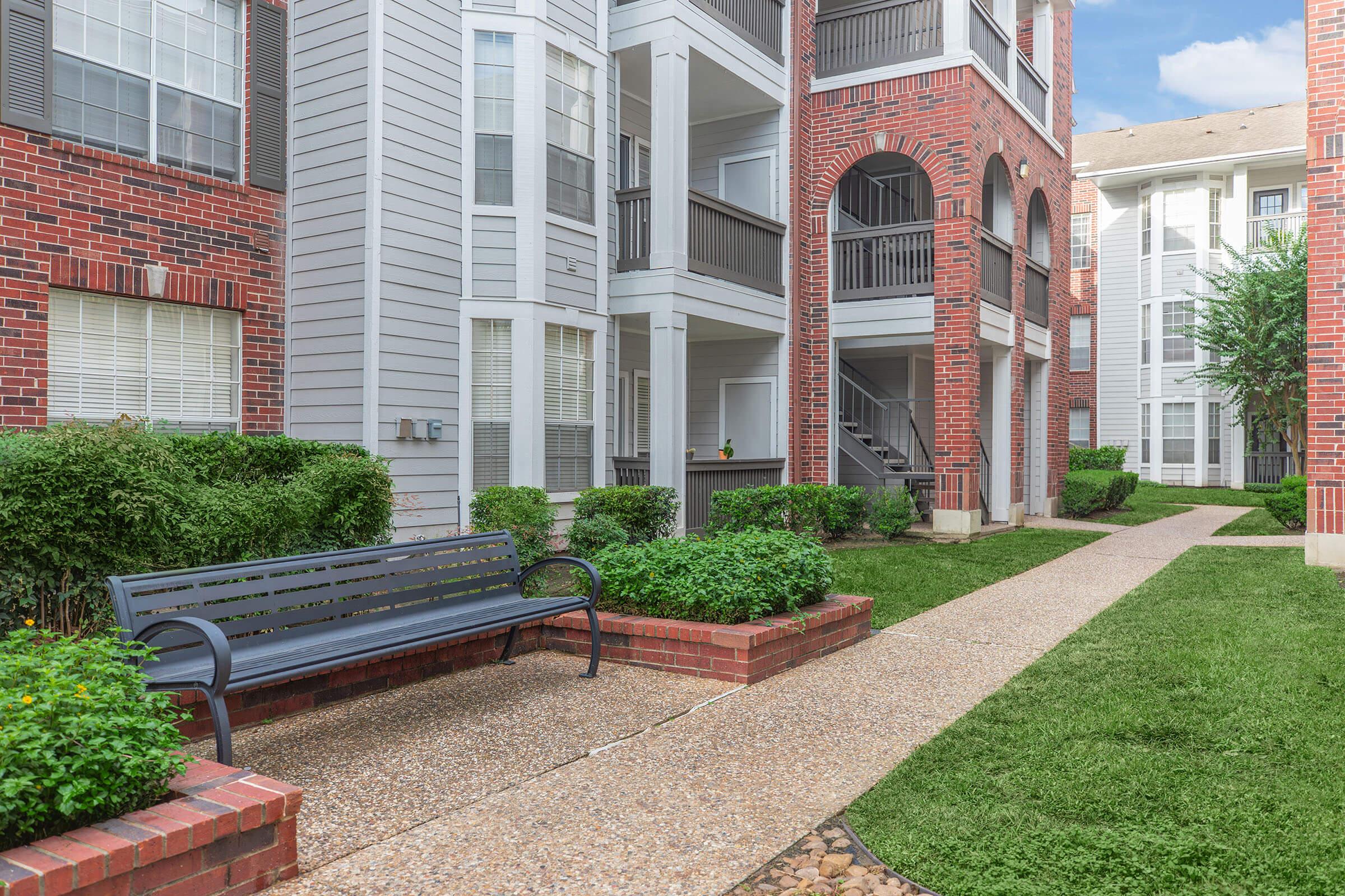 A pathway through a landscaped courtyard featuring greenery, a bench for seating, and two multi-story apartment buildings constructed with a mix of brick and gray siding. The scene is bright, showcasing a well-maintained outdoor living space.
