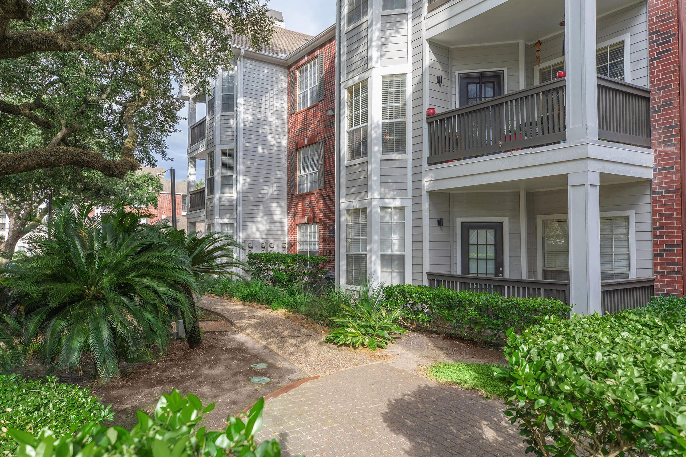 A peaceful residential area featuring an apartment building with a mix of grey and red facades. The scene includes lush greenery, shrubs, and a winding pathway leading to the entrance, set under a partly cloudy sky.