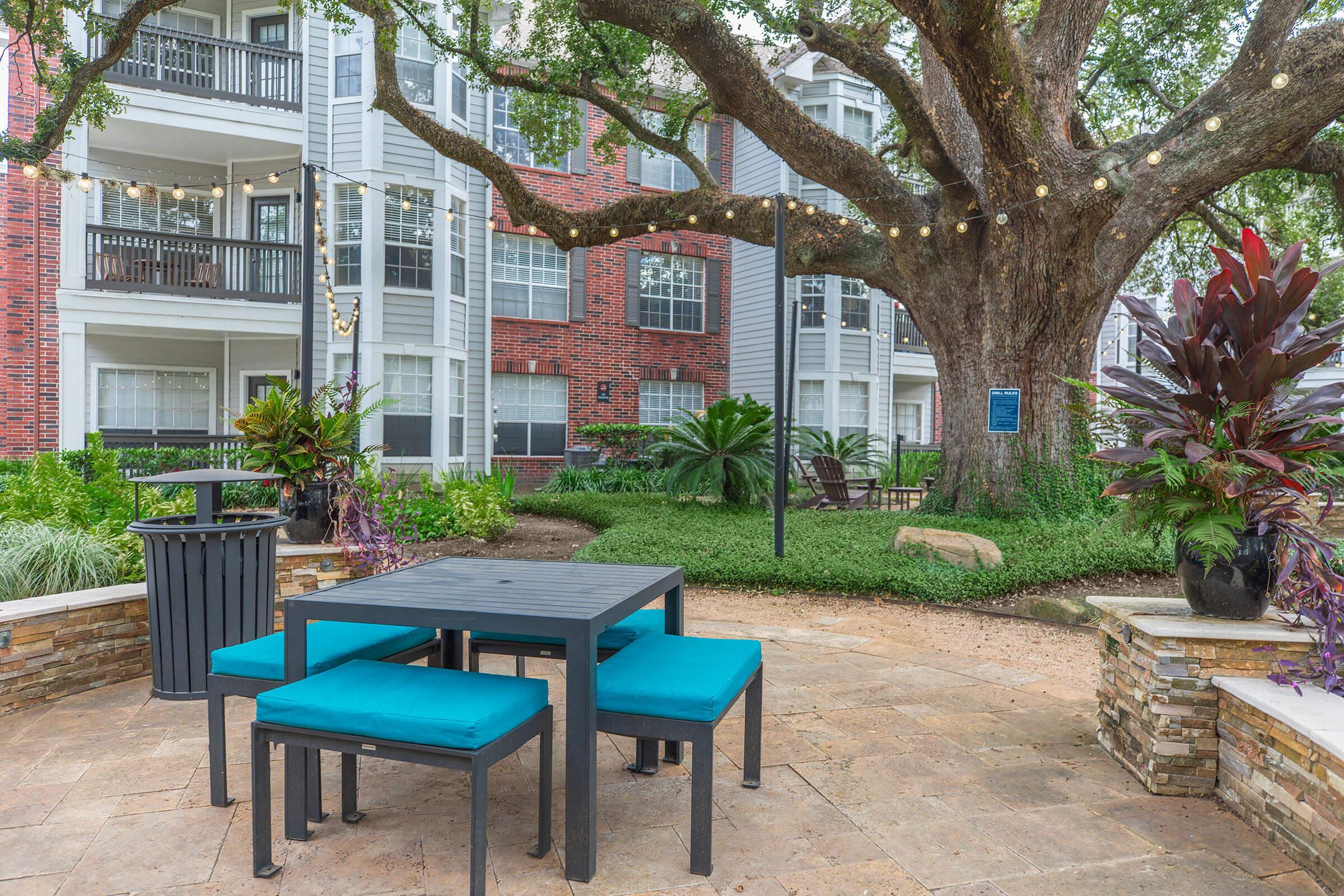 A cozy outdoor seating area featuring a dark wooden table surrounded by turquoise chairs, set amidst lush greenery and decorative plants. Above, string lights hang from a large tree, creating a warm and inviting atmosphere near residential buildings.