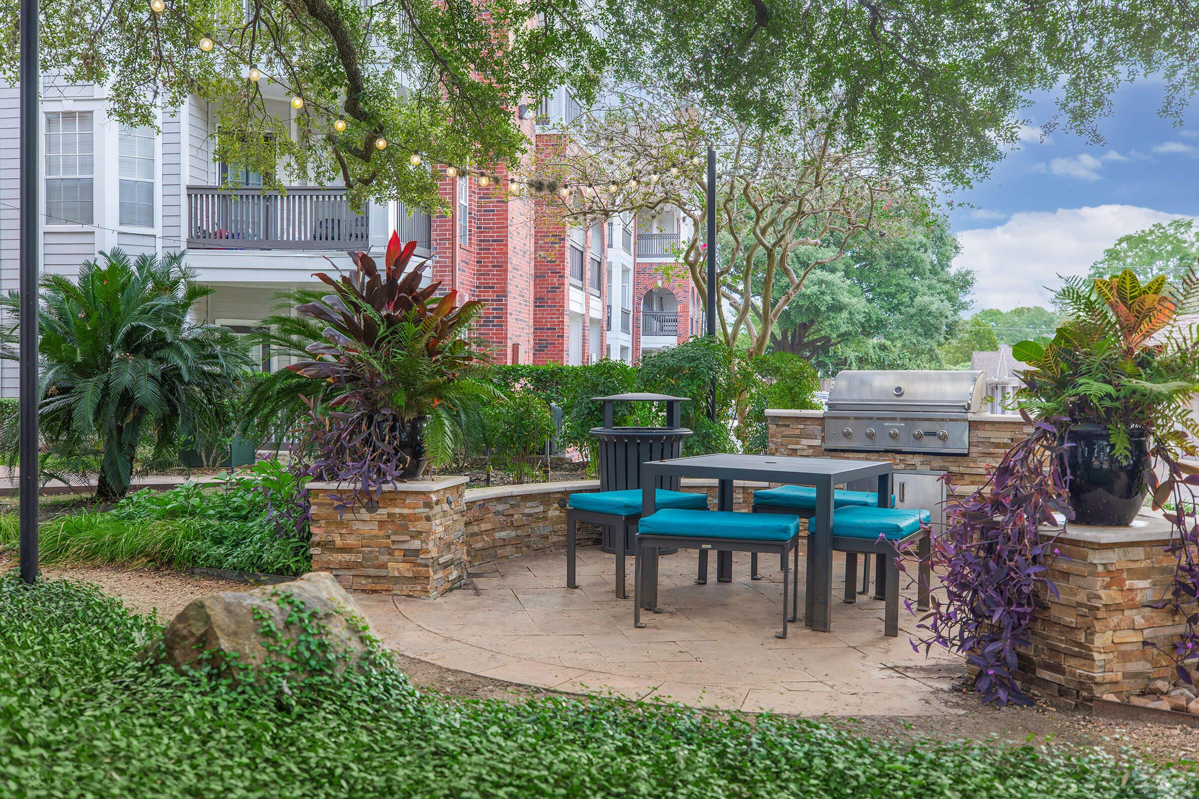 Outdoor patio area featuring a stone seating arrangement with turquoise cushions, a grill, and lush greenery, including various plants and trees, set against a bright blue sky. String lights add a decorative touch to the space, creating an inviting atmosphere for gatherings.