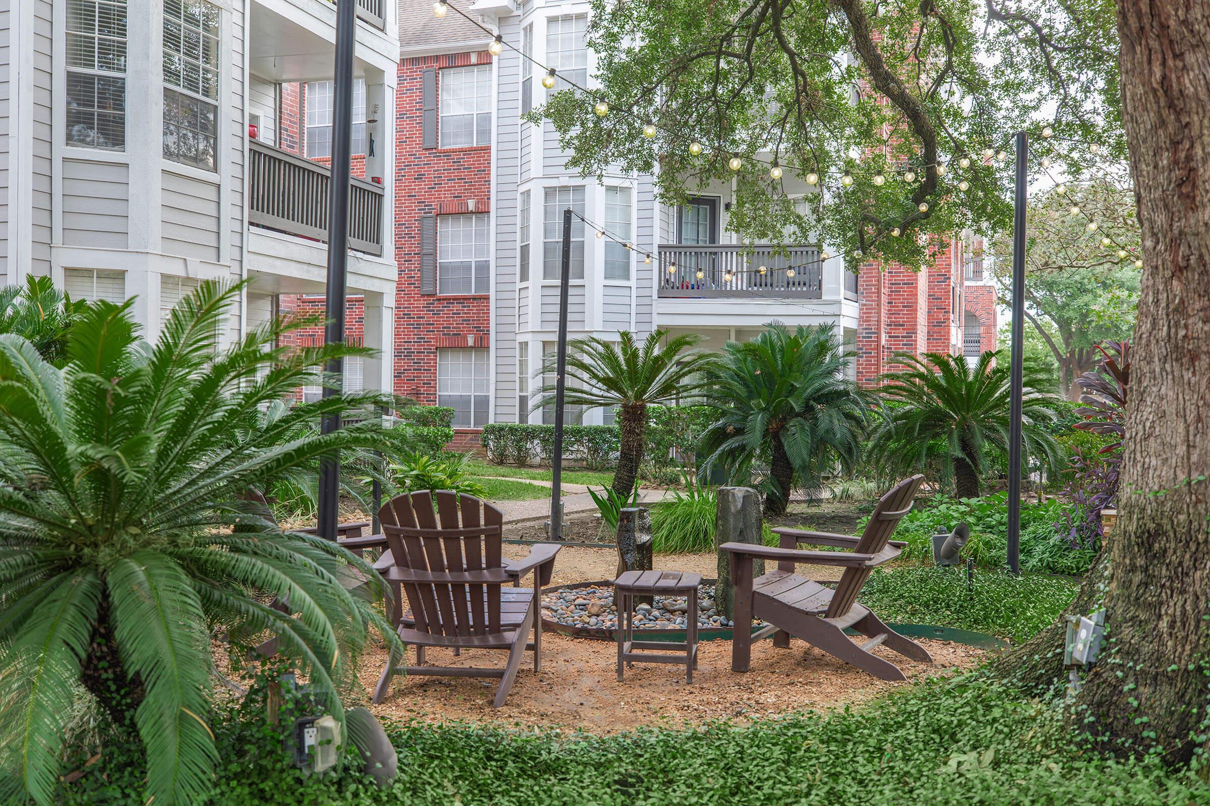 A serene outdoor seating area featuring two wooden Adirondack chairs surrounded by lush greenery, including palm trees and ferns. The background shows a multi-story apartment building with balconies and string lights above. A small fire pit sits at the center of the seating arrangement, on a sandy ground.