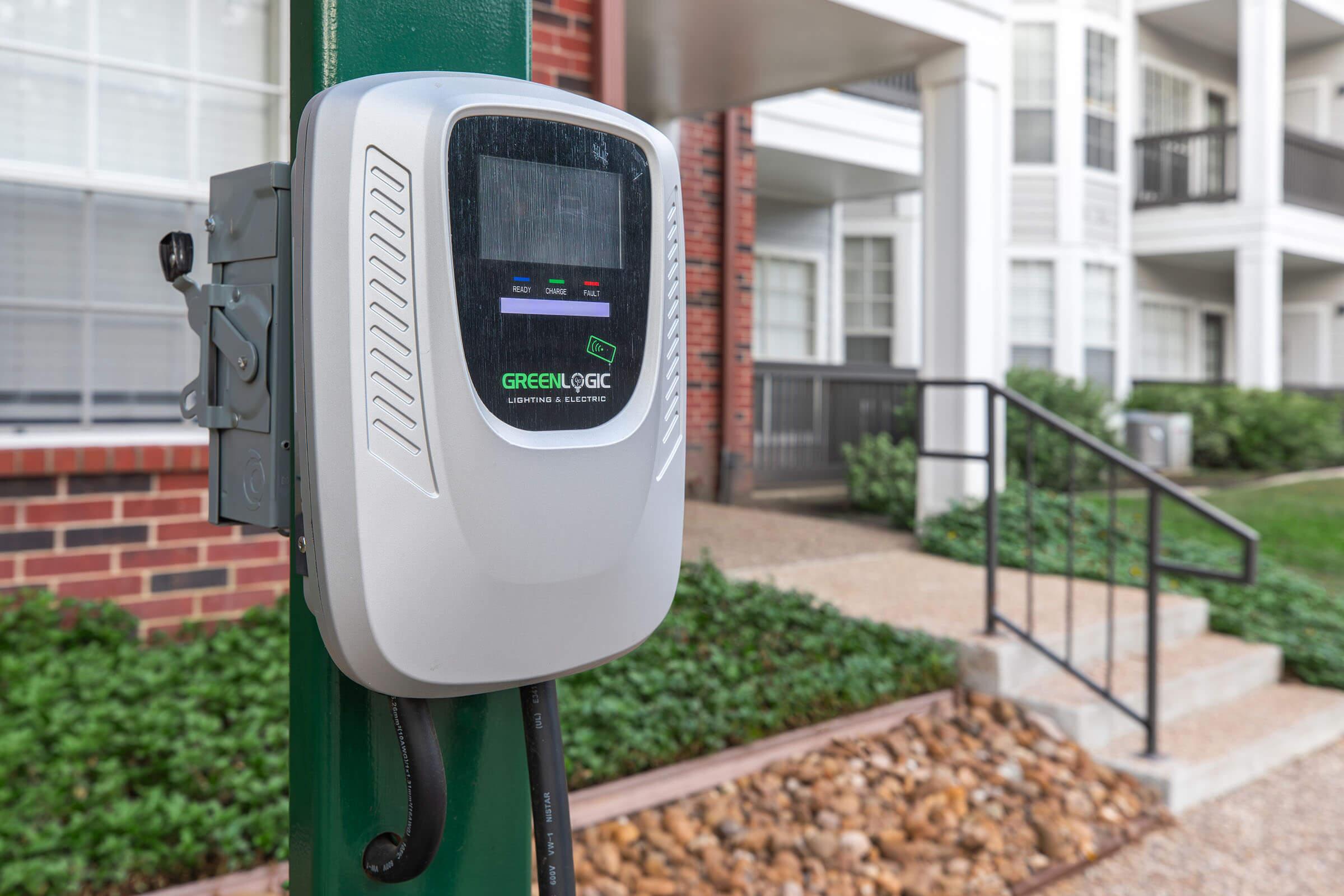 A close-up of a green and gray electric vehicle charging station mounted on a pole, with a digital display showing charging status. In the background, a modern apartment building with multiple balconies and a landscaped pathway is visible. The area features decorative stone landscaping.