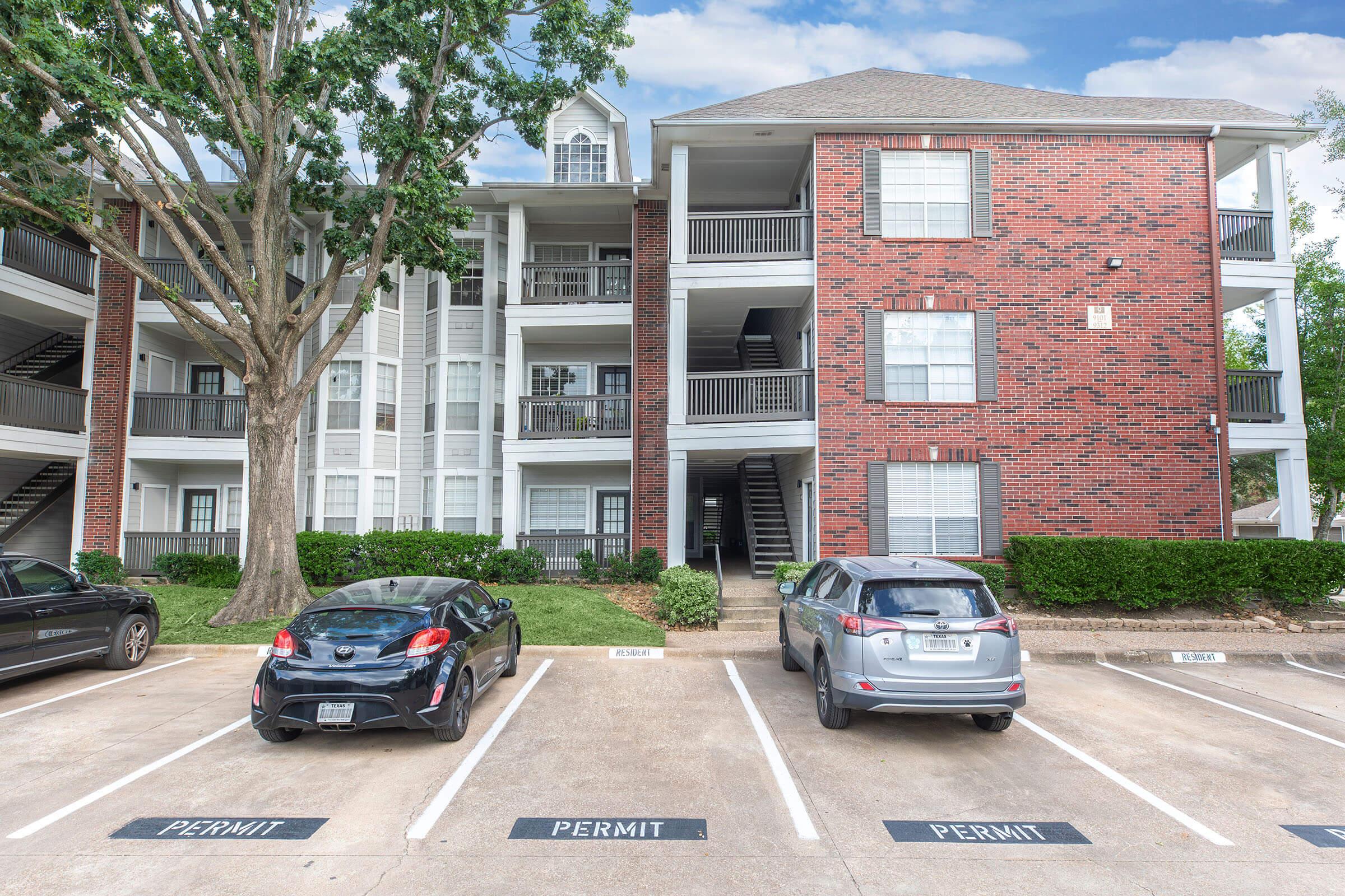 A multi-story apartment building with red brick and white siding, featuring balconies. Two cars parked in front, marked with "PERMIT" signs. A large tree is visible on the left, and there are several green bushes in front of the building. Clear blue sky with scattered clouds above.