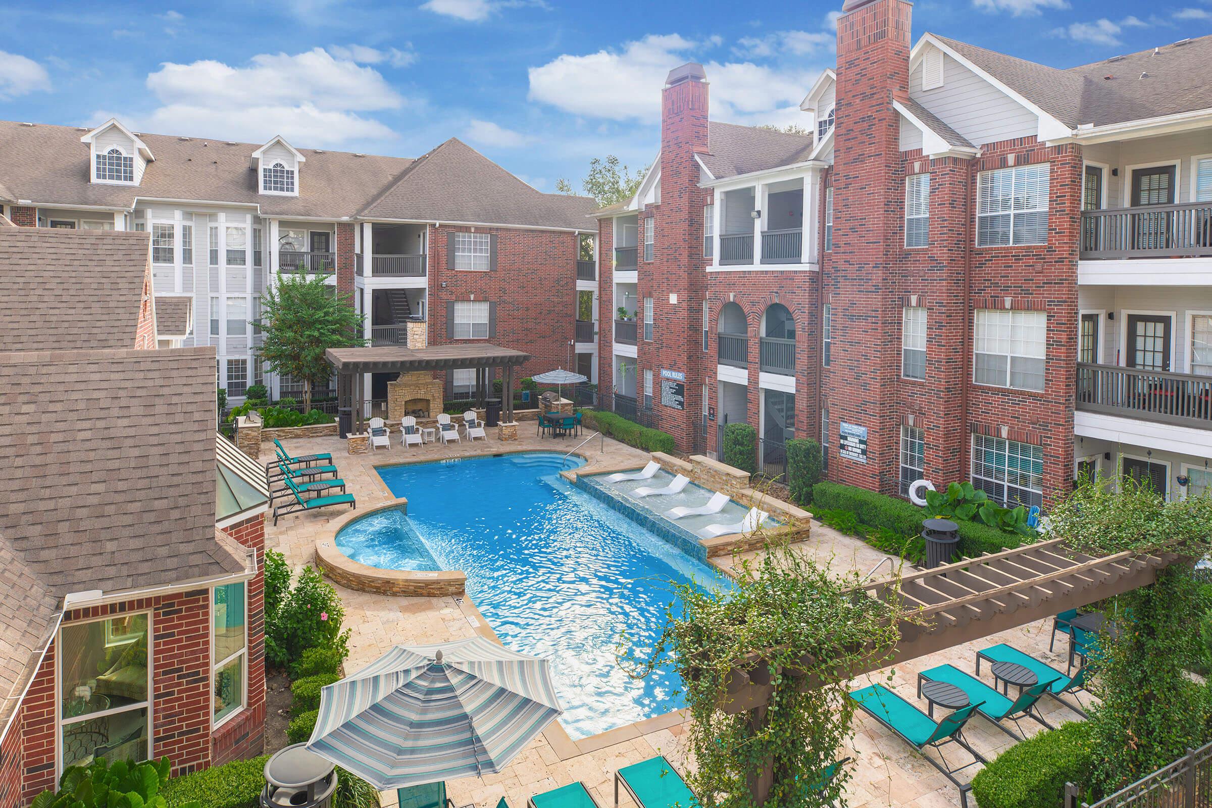 Aerial view of a suburban apartment complex featuring a large blue swimming pool surrounded by lounge chairs and umbrellas. Brick buildings are visible in the background, with greenery and landscaping enhancing the outdoor space. The sky is clear with a few clouds.