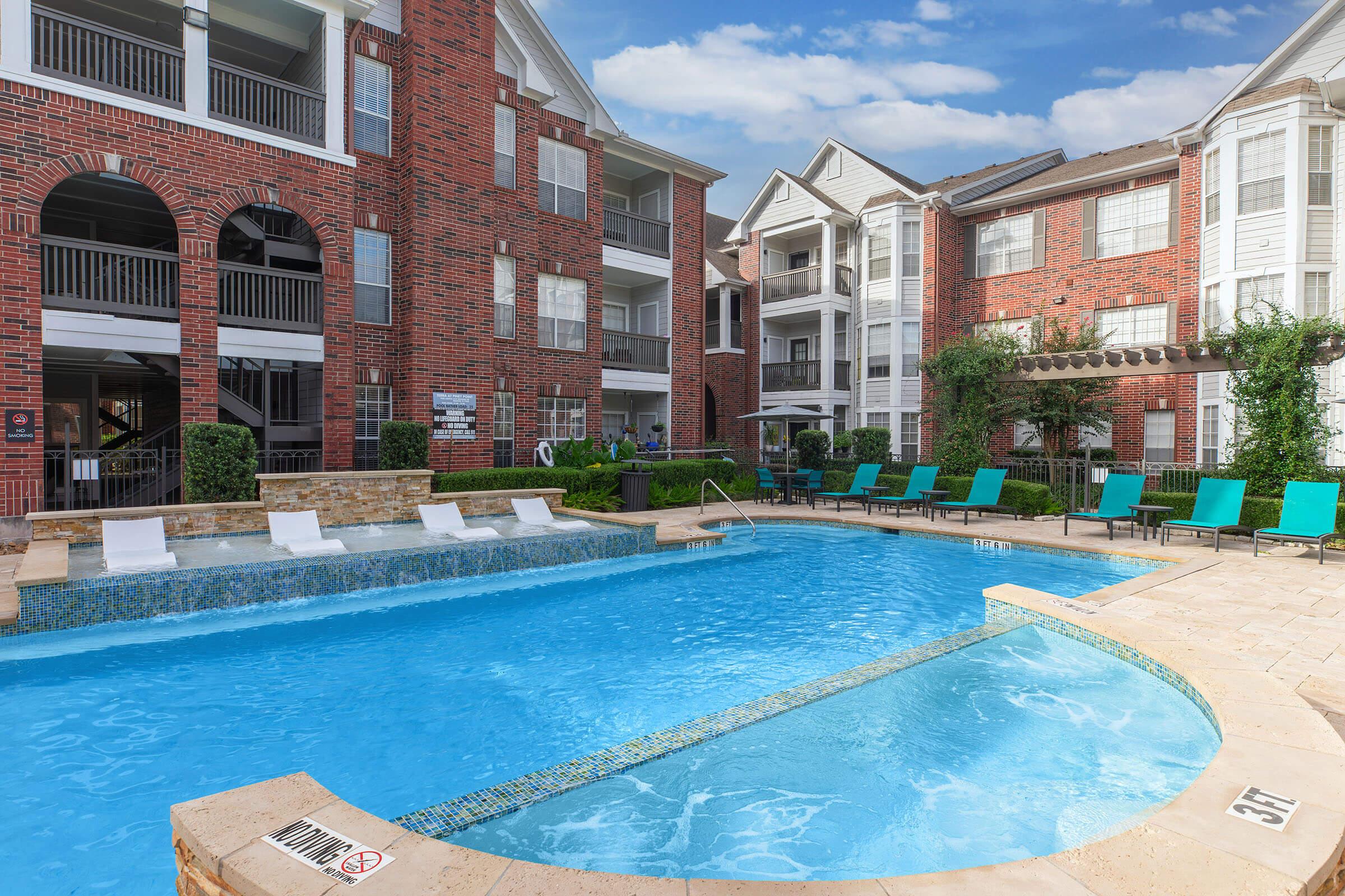 A vibrant apartment complex featuring a swimming pool surrounded by lush landscaping. The pool area includes white lounge chairs and green poolside seating. Several buildings with brick exteriors are visible in the background under a blue sky.