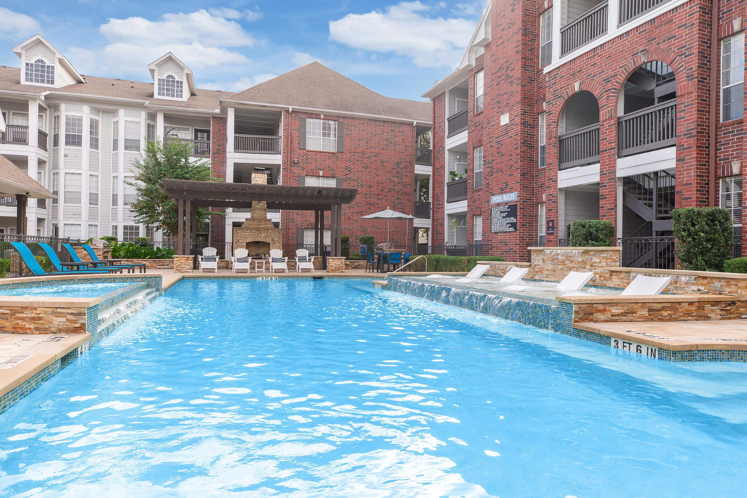 A large swimming pool in a well-maintained apartment complex. The pool features a shallow section with lounge chairs and a stone fire pit nearby, surrounded by brick buildings and lush greenery. Clear blue sky and a few clouds enhance the serene atmosphere.