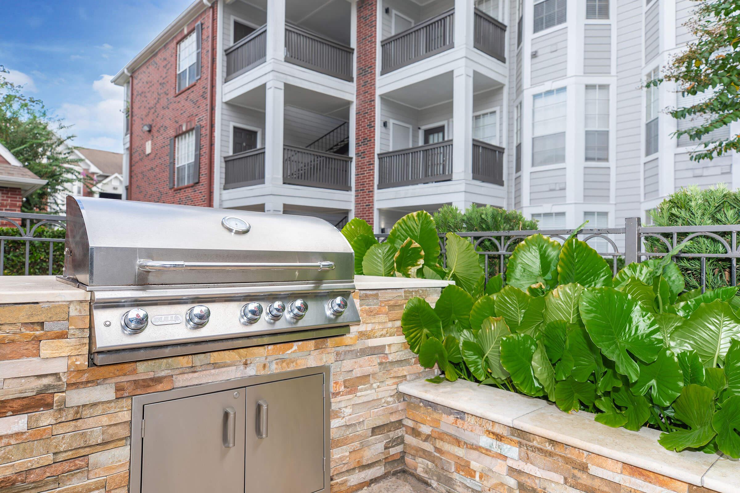 Outdoor grill station with a stainless steel barbecue on a stone surface, surrounded by lush green foliage and a view of apartment balconies in the background. The setting is well-maintained and inviting, perfect for gatherings or outdoor cooking.