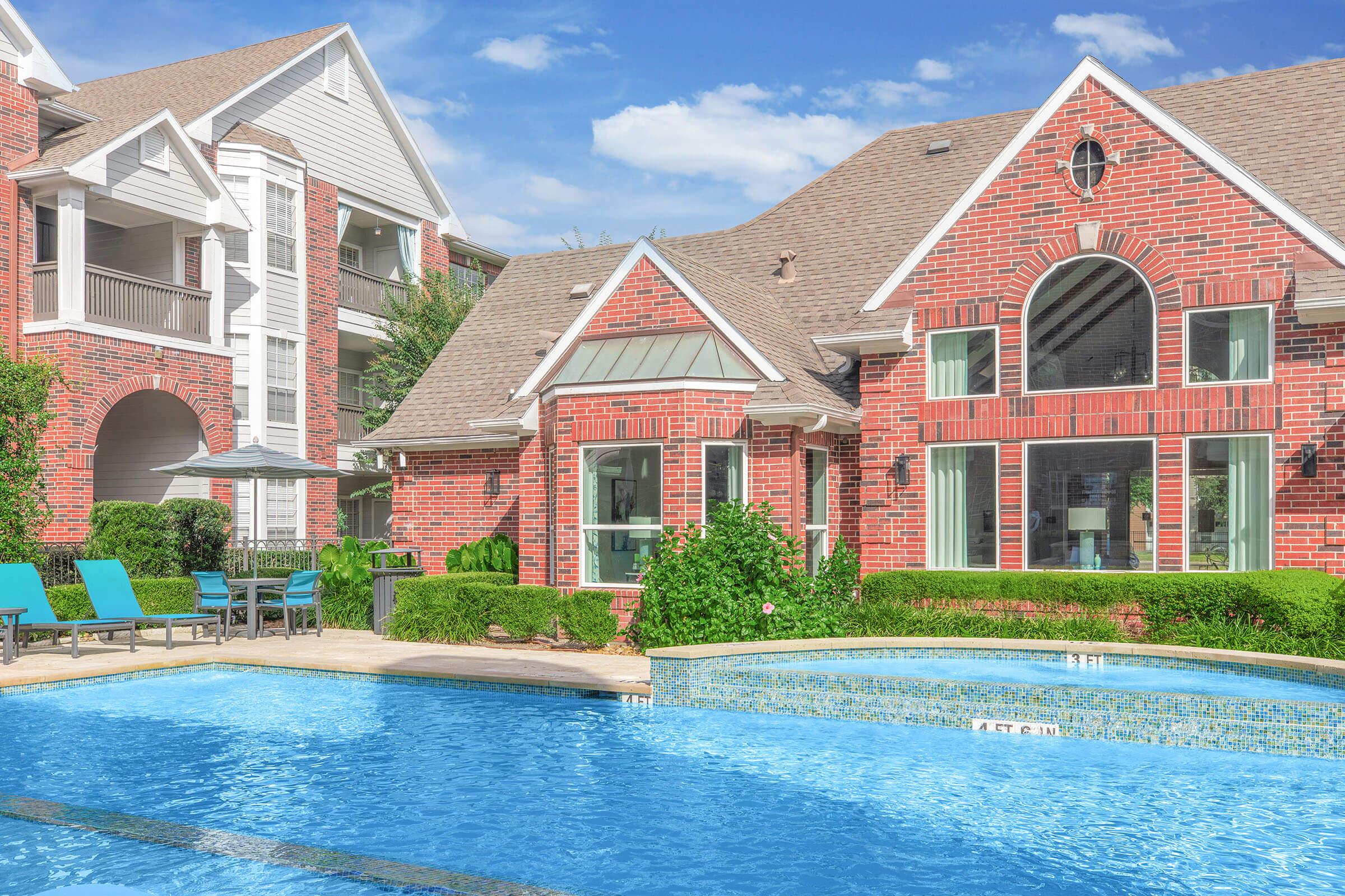 A view of a luxurious residential area featuring a brick exterior house beside a sparkling blue swimming pool. Surrounding the pool are lounge chairs and neatly manicured greenery. In the background, additional apartment buildings can be seen under a clear blue sky.