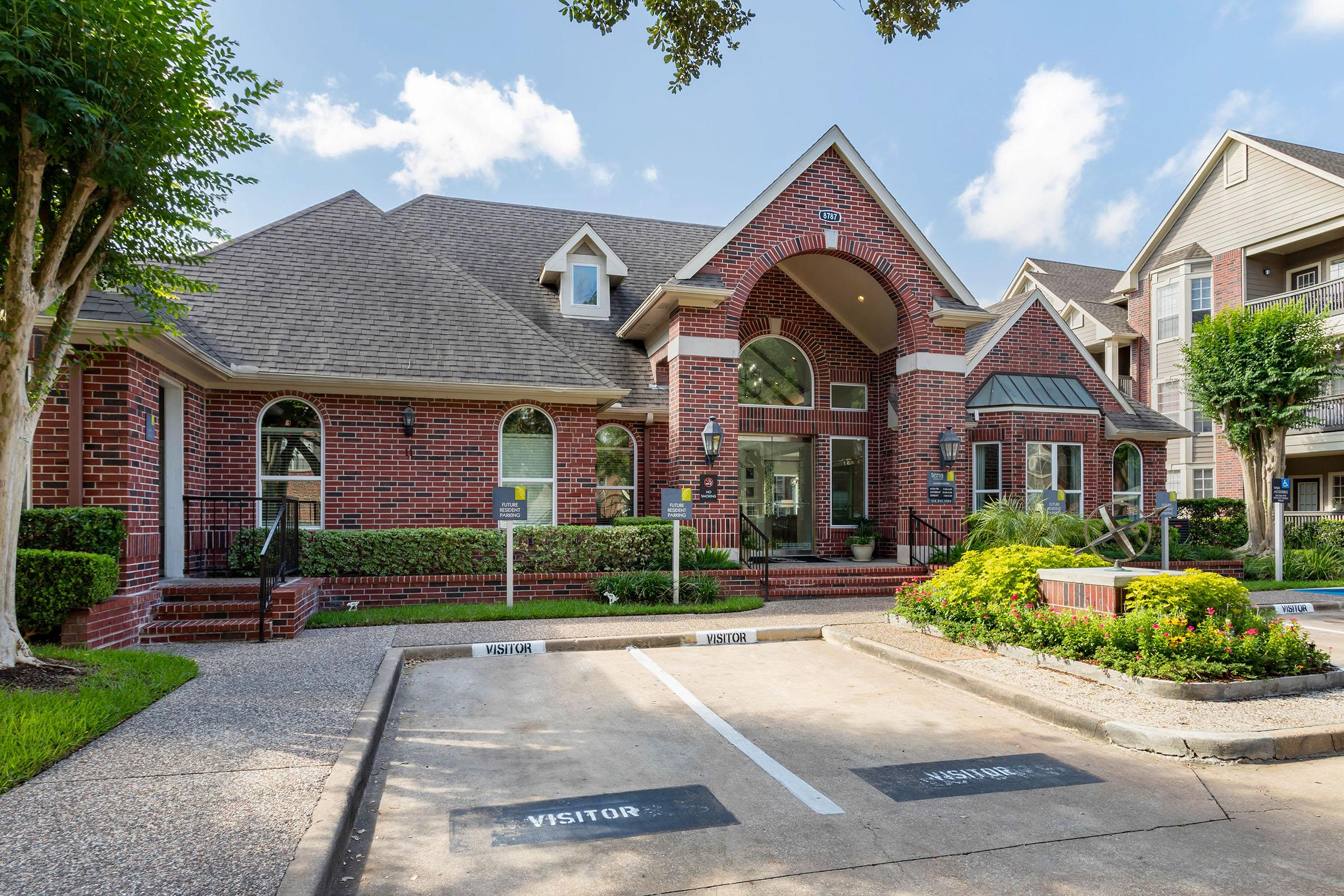 A brick building featuring an arched entrance, surrounded by trees and landscaped greenery. The parking area displays "Visitor" signs. Blue skies with few clouds are visible above, indicating a bright day. The architecture suggests a welcoming atmosphere, likely for an apartment complex or community center.
