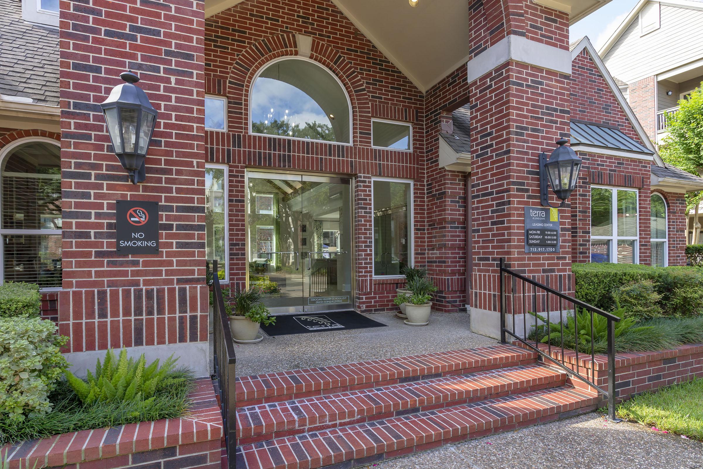 Exterior view of a brick building entrance featuring large windows, a "No Smoking" sign, and decorative lighting. The pathway is framed by potted plants and well-maintained landscaping, leading to double glass doors.