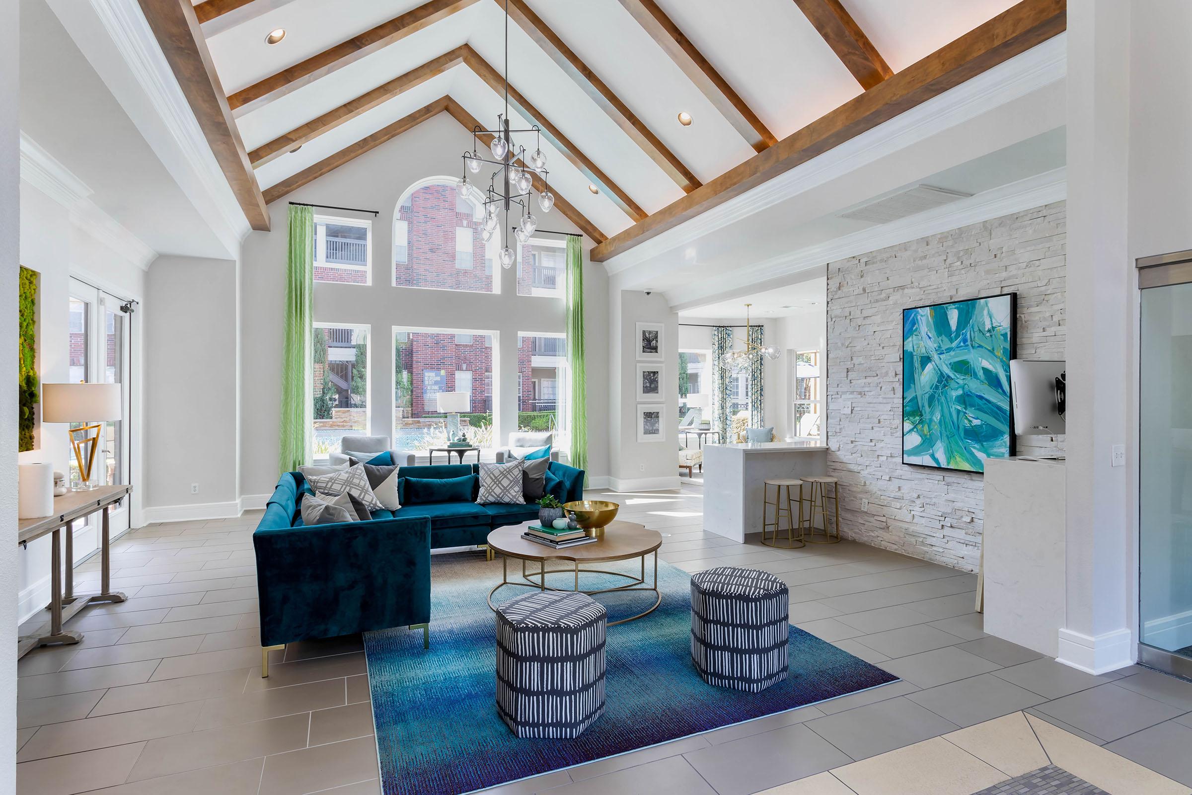 A modern living room featuring a high vaulted ceiling with wooden beams, large windows letting in natural light, a teal sectional sofa, a round coffee table, and two patterned poufs. There’s a stone accent wall and decorative artwork nearby, creating a stylish and inviting atmosphere.