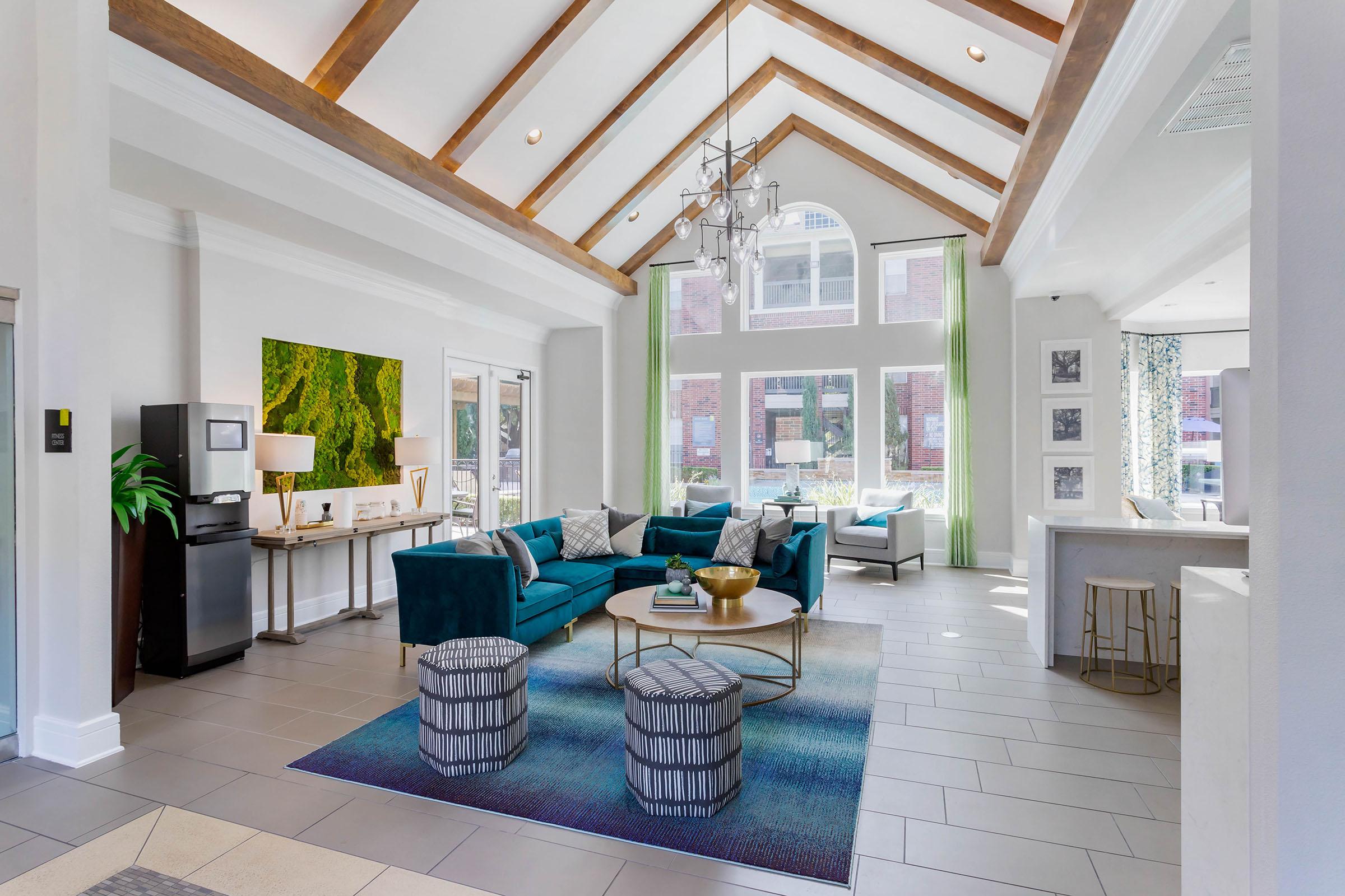 A bright and spacious living room with high vaulted ceilings and wooden beams. The room features a teal sofa, decorative pillows, and two patterned ottomans on a textured rug. Large windows allow natural light in, and there's a modern dining area visible in the background. Green curtains add a pop of color.