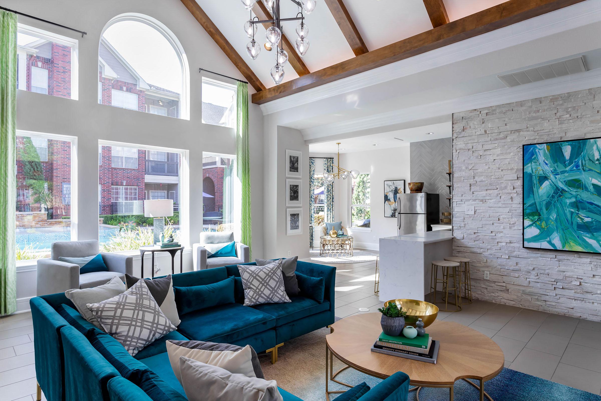 A modern living room featuring a high ceiling with wooden beams, large windows with green curtains, and stylish blue sofas adorned with decorative pillows. A round coffee table sits in the center, and a cozy dining area is visible in the background. The decor includes artwork and a light color palette, creating an inviting atmosphere.