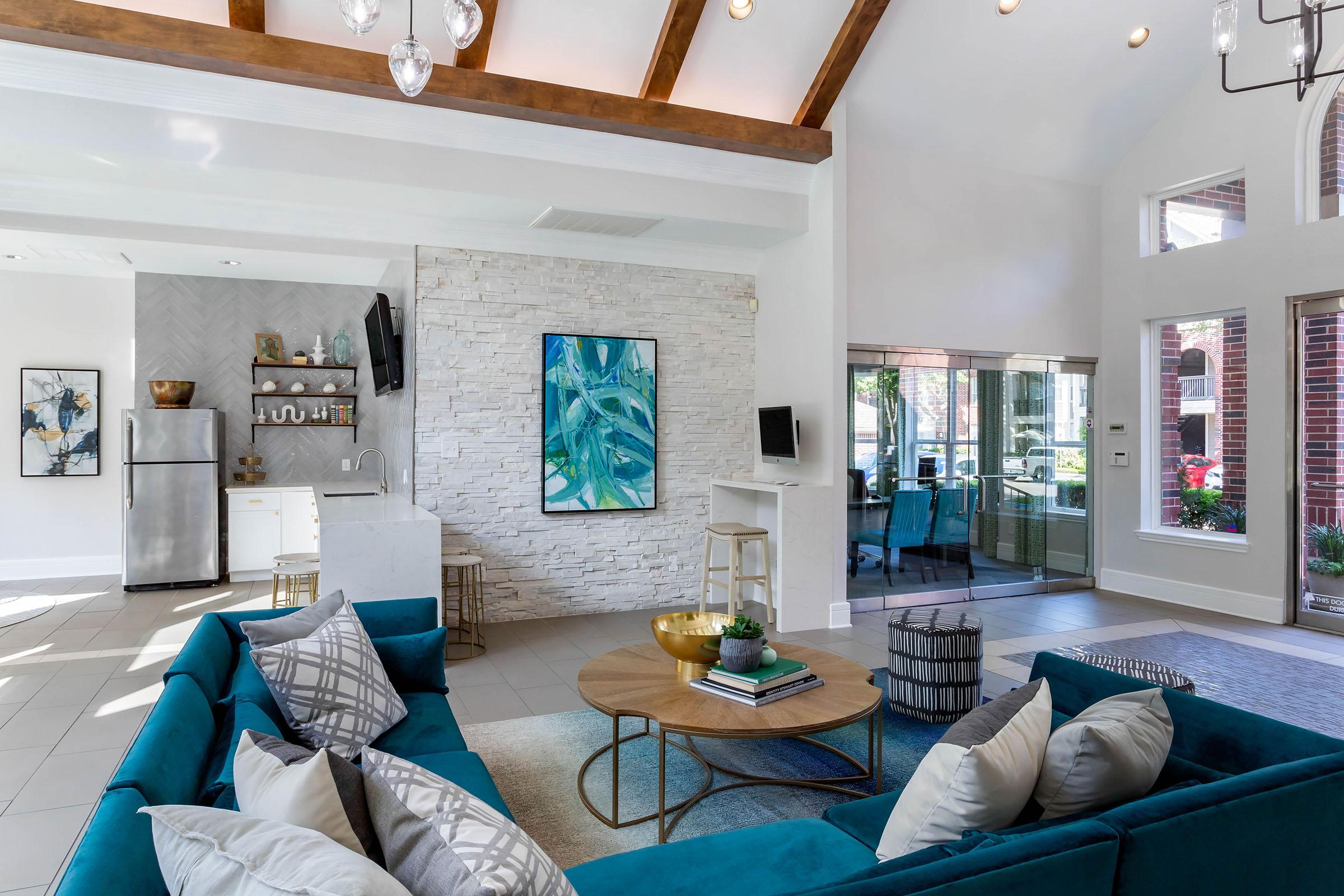 A modern living room featuring a teal sectional sofa with decorative pillows, a round coffee table on a light-colored rug, and a stone accent wall. Sunlight streams in through large windows, highlighting a stylish kitchen area in the background and a piece of abstract art on the wall.