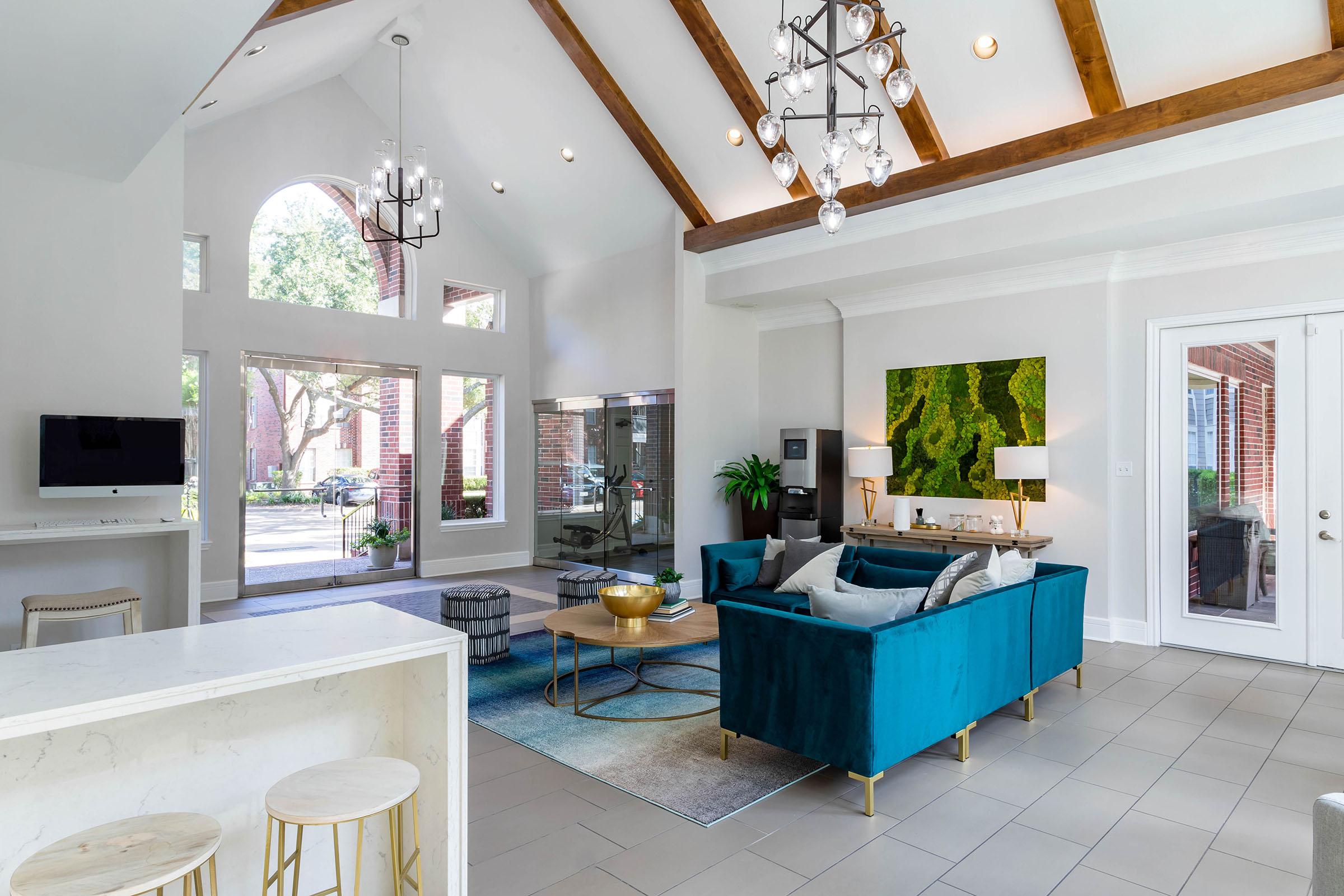 A modern living room featuring high ceilings with wooden beams, large windows allowing natural light, and stylish furnishings including a blue velvet sofa, a gold coffee table, and decorative plants. A sleek TV area and a contemporary artwork add to the inviting ambiance.