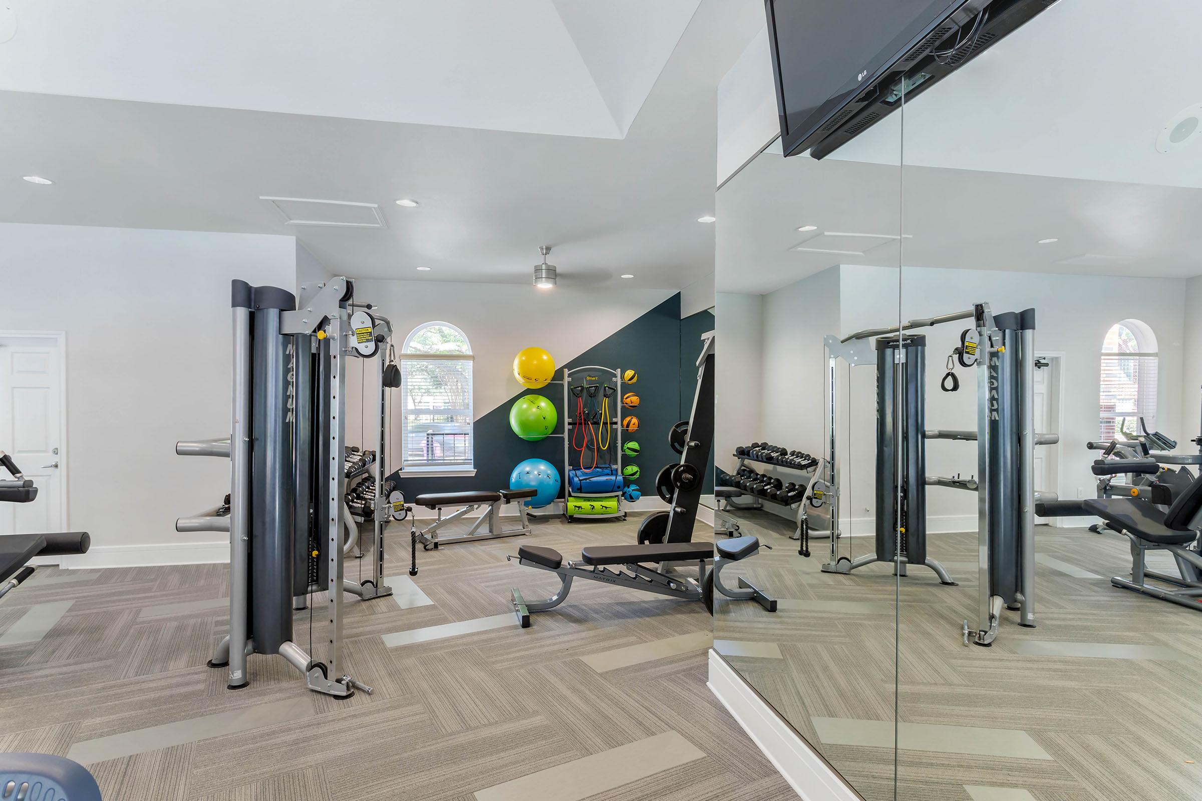 A modern gym interior featuring various exercise equipment, including weight machines, free weights, and exercise balls. Large mirrors reflect the space, enhancing the feeling of openness. Natural light enters through windows, creating a bright and inviting atmosphere for workouts.