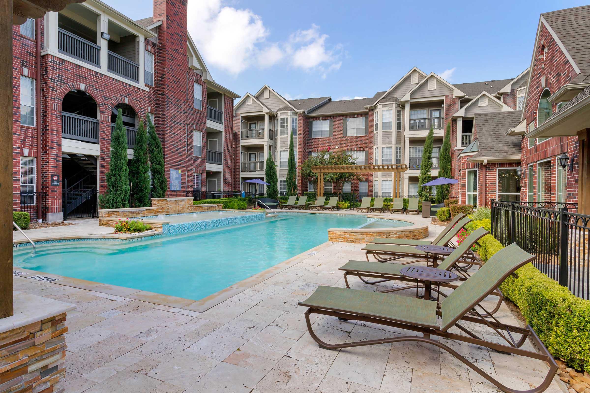 A serene outdoor pool area surrounded by brick apartment buildings. Lounge chairs are arranged along the edge of the pool, with lush green landscaping and tall trees adding to the ambiance. Clear blue skies and fluffy clouds complete the relaxing atmosphere of this residential complex.