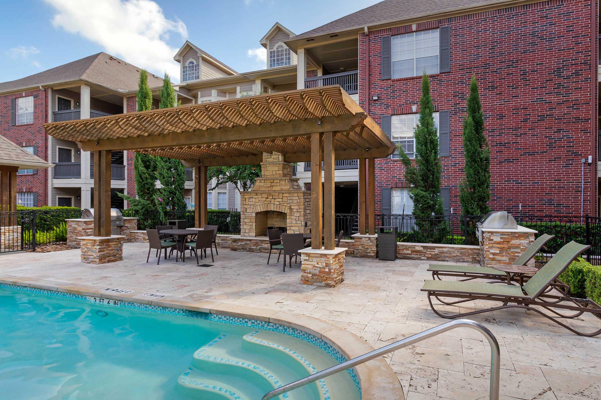 A relaxing outdoor area by a swimming pool featuring a wooden pergola with a stone fireplace, surrounded by lawn, lounge chairs, and tall leafy plants, with multi-story brick apartment buildings in the background.