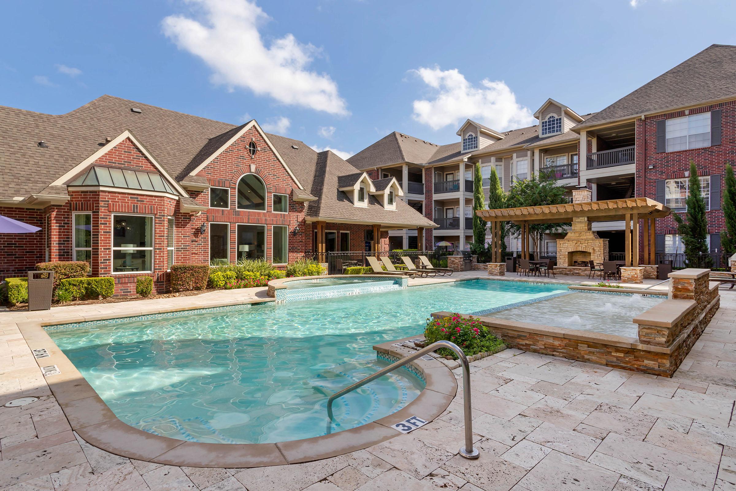 A modern outdoor swimming pool area surrounded by a stone patio, lounge chairs, and a decorative fireplace. The background features a stylish brick building with multiple balconies and lush greenery under a partly cloudy blue sky.