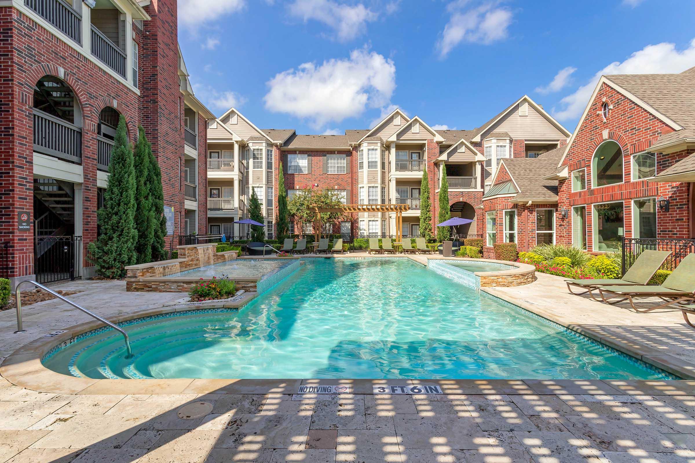 A well-maintained outdoor swimming pool surrounded by landscaped gardens and lounge chairs, situated in a modern apartment complex. The buildings feature brick exteriors and balconies, with clear blue skies providing a bright atmosphere.