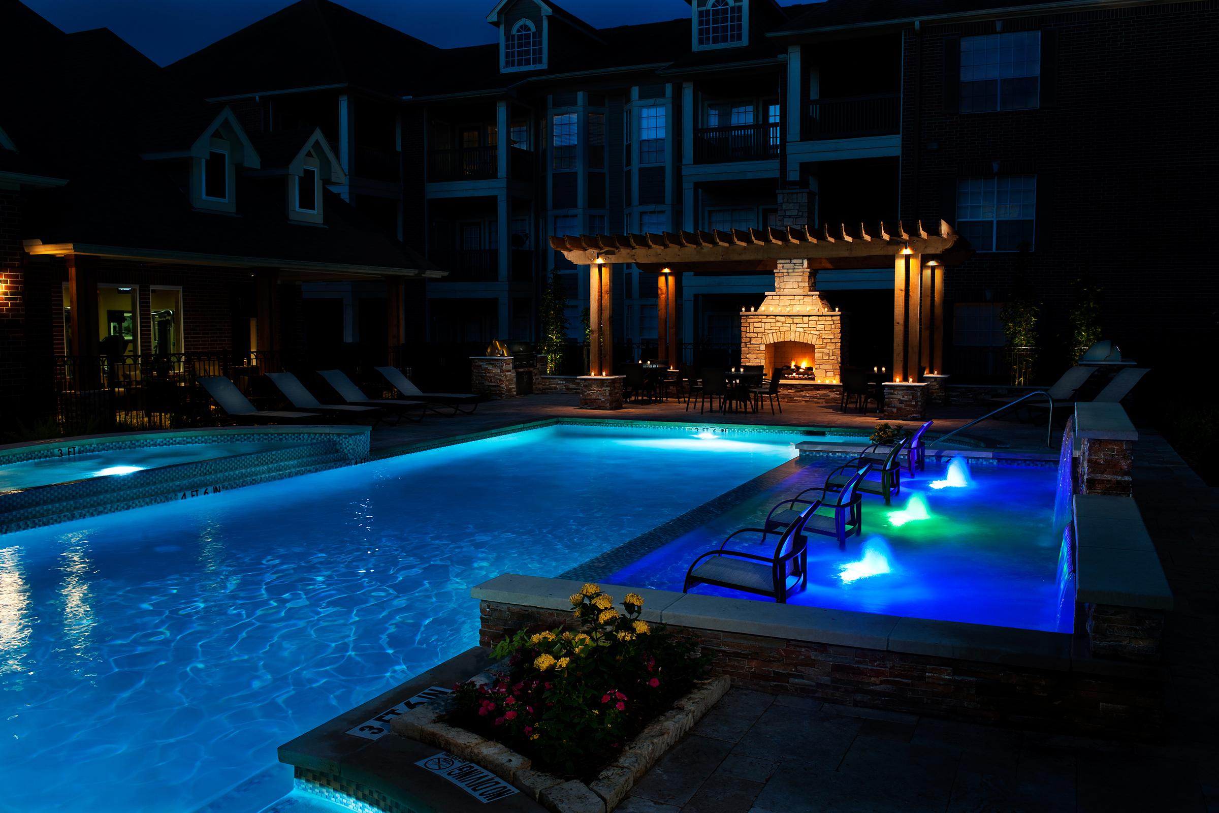 A beautifully illuminated outdoor pool area at night, featuring a sparkling blue pool with colorful lights, lounge chairs, and a cozy fireplace under a pergola. Surrounding plants and flowers add a touch of nature to the serene atmosphere of the apartment complex.