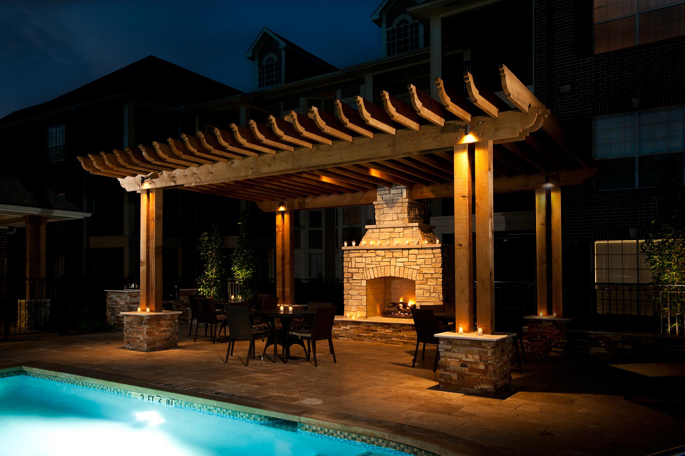 A beautifully lit outdoor patio with a wooden pergola, featuring a stone fireplace and a dining area with chairs around a table. The setting is near a swimming pool, surrounded by soft landscaping, creating a cozy ambiance at night.