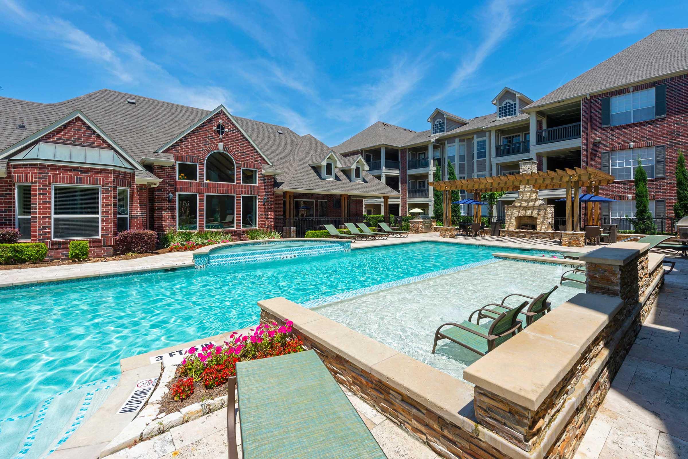 A well-maintained apartment complex pool area featuring a crystal-clear swimming pool surrounded by lounge chairs, vibrant flower beds, and brick buildings under a clear blue sky. Elegant outdoor seating and a decorative pergola enhance the inviting atmosphere.