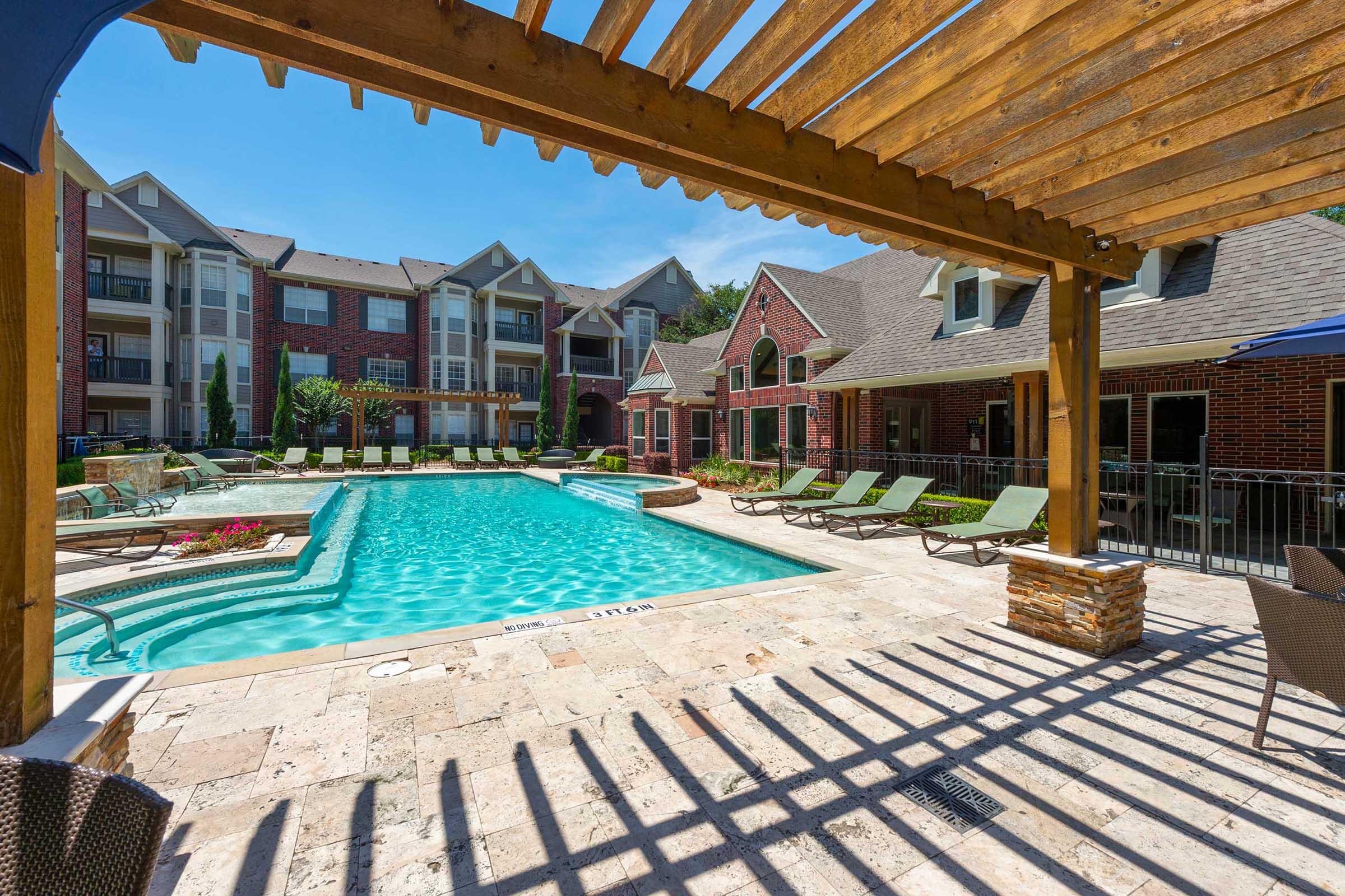 A luxurious outdoor swimming pool surrounded by lounge chairs, set in a beautifully landscaped apartment complex. A wooden pergola provides shade, and the bright blue sky enhances the inviting atmosphere. The pool area features stone accents and is bordered by well-maintained greenery and buildings.