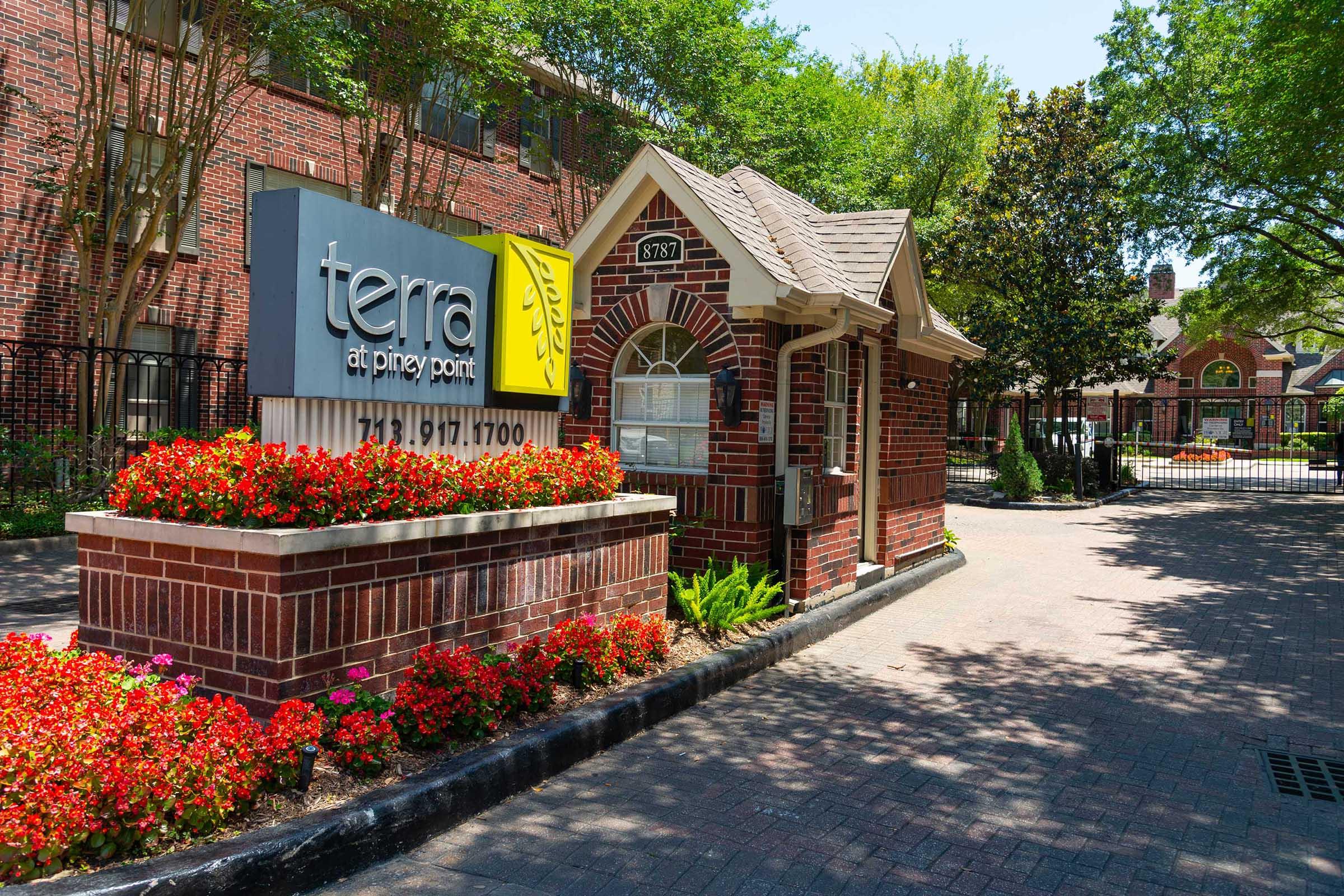 Brick entrance with a sign for "terra at pinery point" featuring flowers in vibrant colors. A small, decorative building is adjacent to the sign, surrounded by greenery and a gated entryway leading to the property. The scene is bright and welcoming.