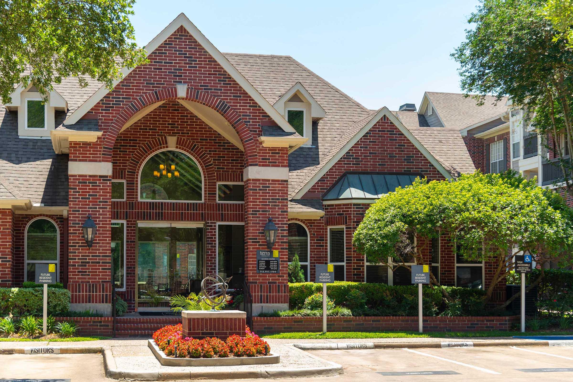 A brick building with an arched entrance, surrounded by lush greenery and colorful flower beds. The exterior features large windows and a well-maintained lawn, suggesting a welcoming atmosphere. A sign is visible near the entrance, indicating it is likely part of an apartment complex or community.