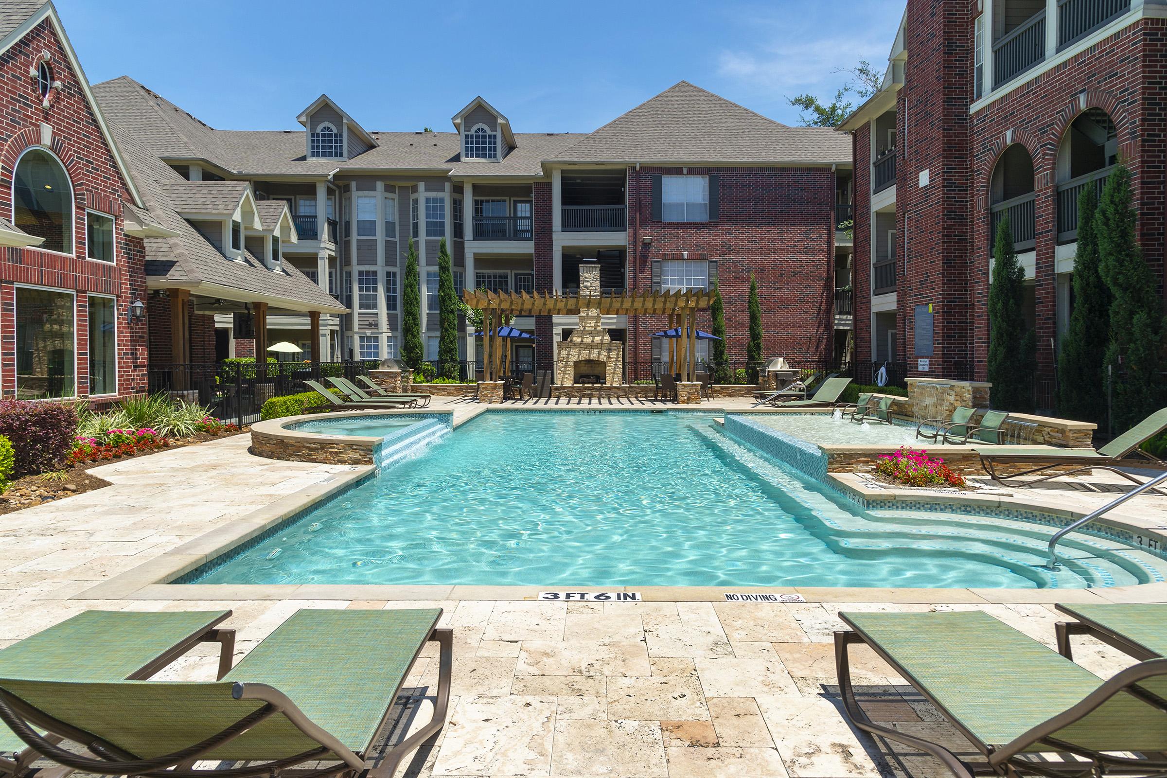 A sunny outdoor pool area featuring a large, clear blue swimming pool surrounded by lounge chairs and colorful landscaping. The pool is bordered by brick buildings with multiple floors. An outdoor fireplace and greenery add to the inviting atmosphere.