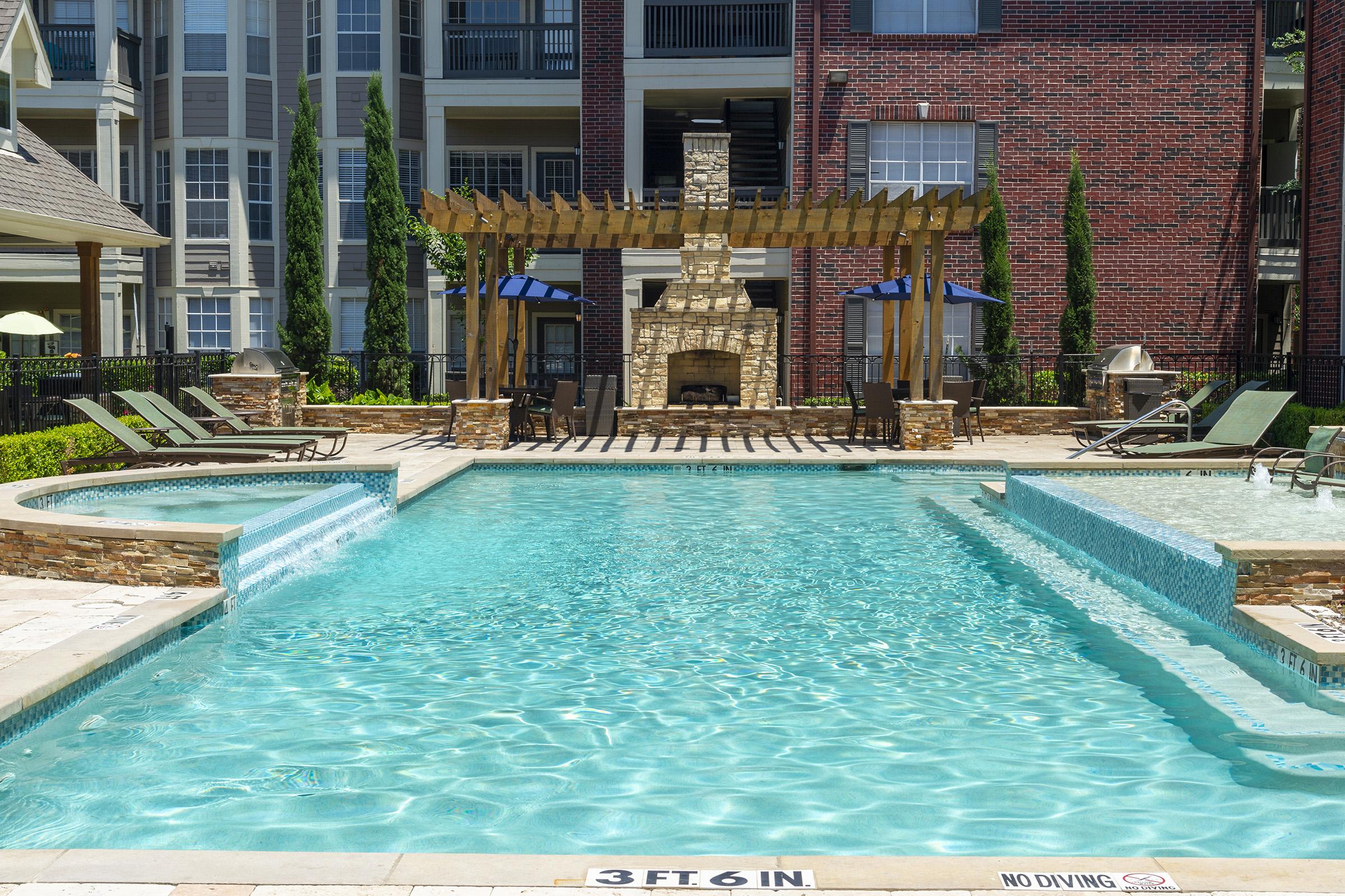 A bright outdoor pool area featuring a large swimming pool with clear blue water, relaxing lounge chairs, and a stone fireplace structure. Lush landscaping surrounds the pool, and a pergola provides shade. The area is in a residential complex with multi-story buildings in the background.