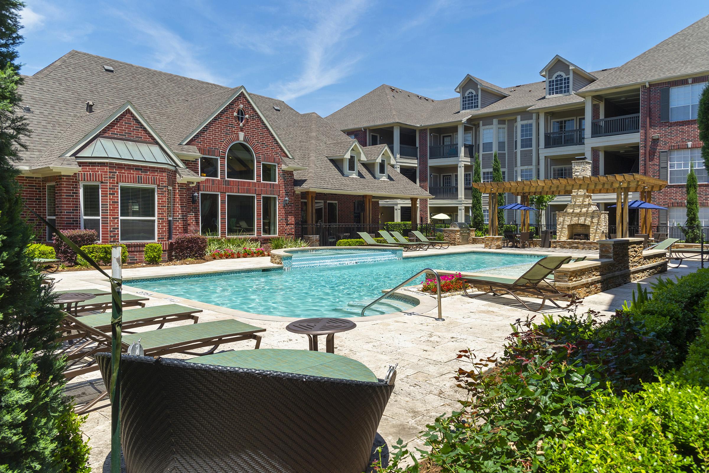 A sunny view of a well-maintained outdoor pool area surrounded by brick buildings and lush landscaping. Lounge chairs are arranged around the pool, and a covered seating area is visible in the background, featuring stonework and greenery. The sky is clear with a few clouds.