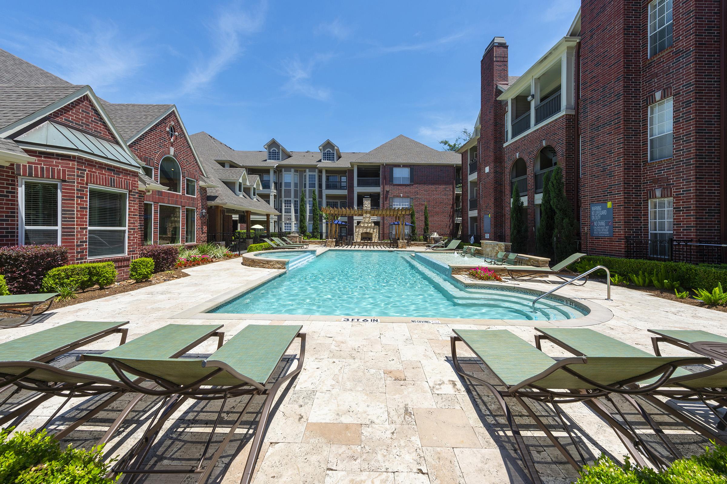 A sunny outdoor pool area featuring a sparkling blue pool surrounded by lounge chairs. The space is landscaped with greenery and flowers, with residential buildings in the background. Clear blue skies enhance the inviting atmosphere of this relaxing setting.