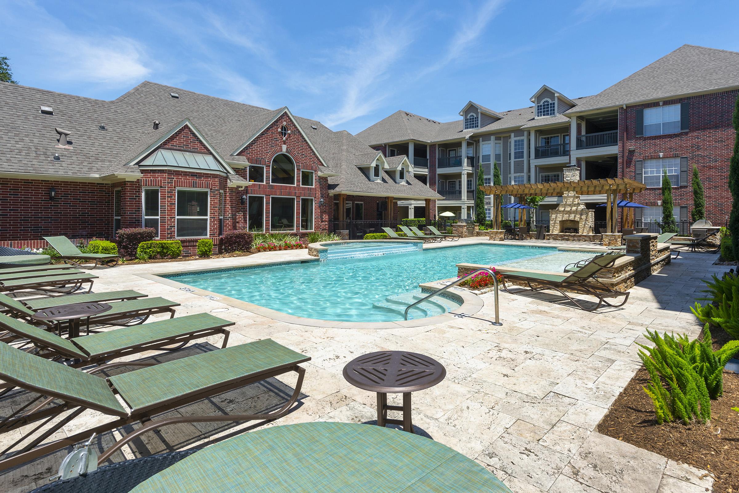 A beautifully designed outdoor pool area surrounded by lounge chairs, nestled among landscaped greenery. The pool features a spa-like design, with stone accents and an outdoor seating area. The backdrop includes a modern multi-story building, showcasing large windows and brick architecture under a clear blue sky.