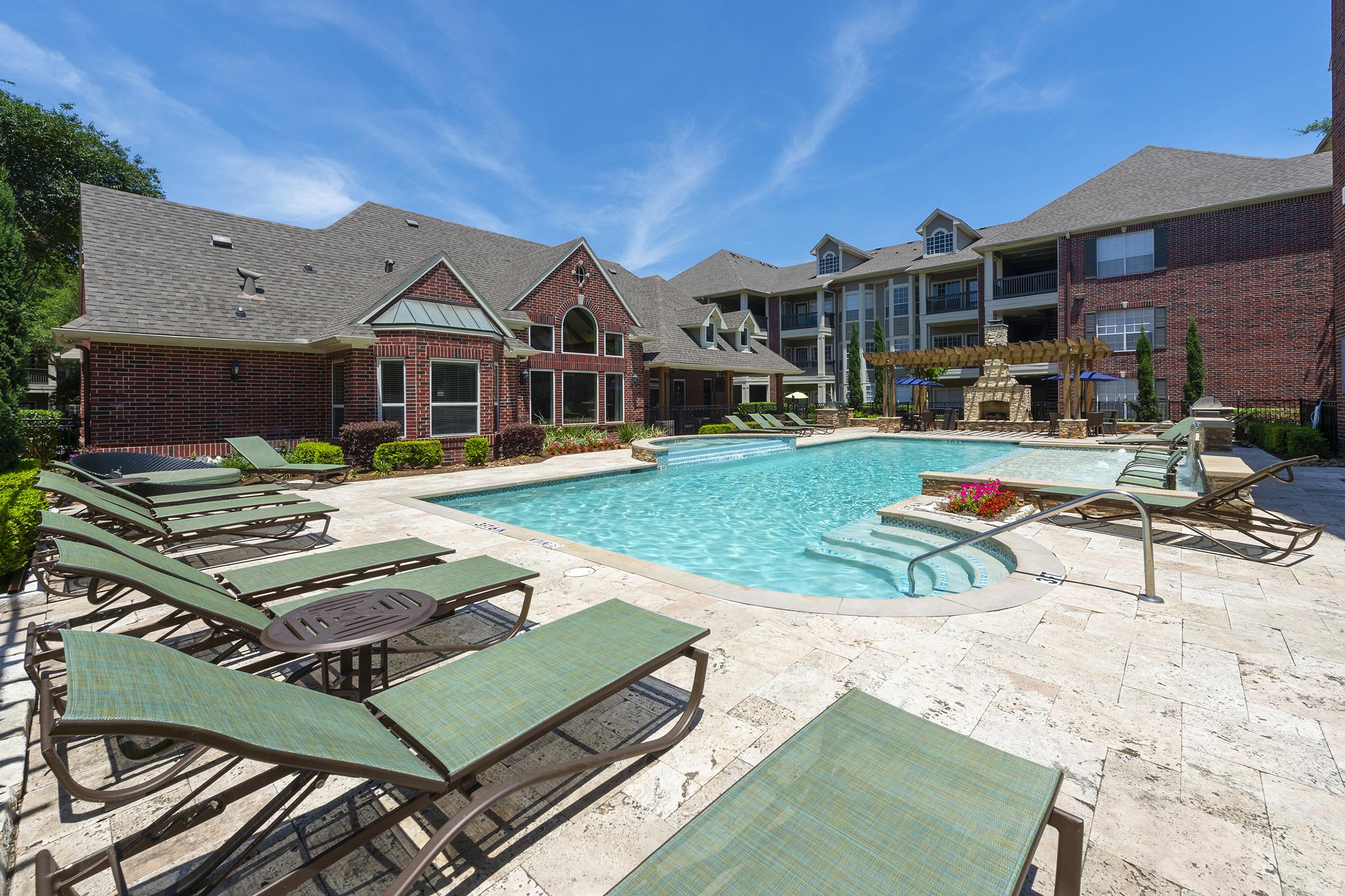 A sunny pool area surrounded by lounge chairs and greenery. In the background, a brick building with a covered patio and outdoor fireplace can be seen. The clear blue sky adds to the inviting atmosphere of the space, ideal for relaxation and leisure.