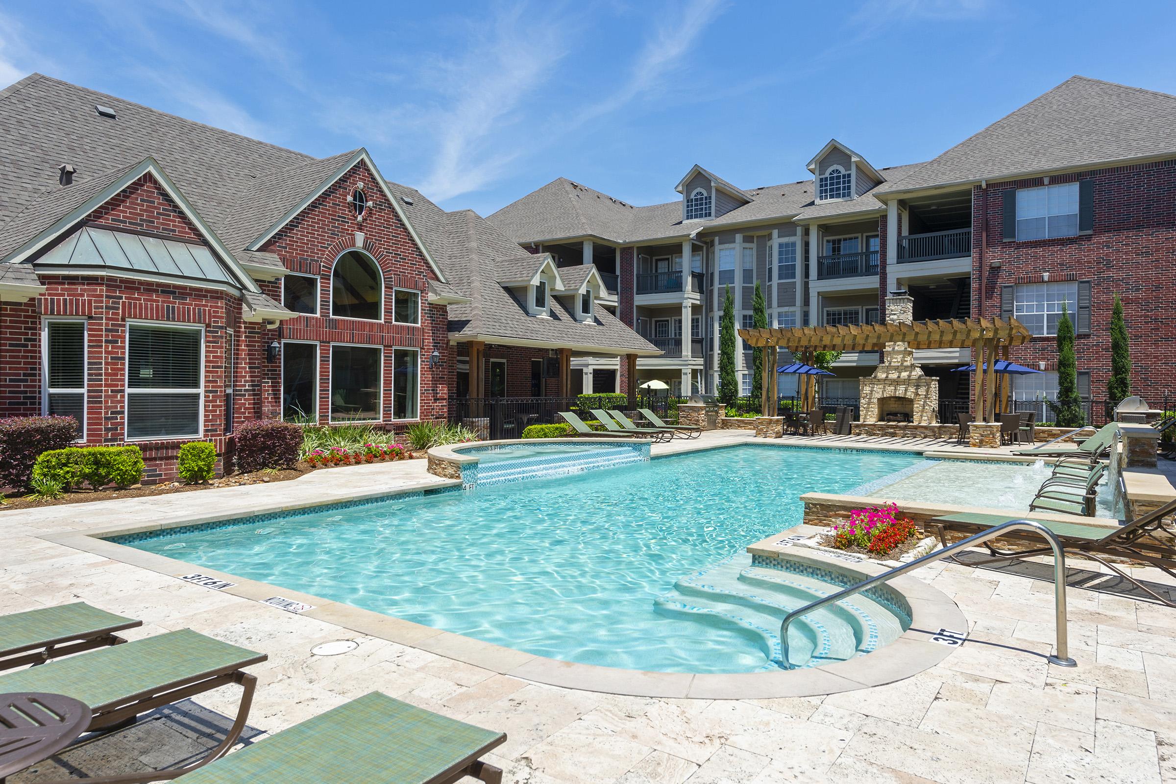 A bright, inviting outdoor pool area surrounded by brick buildings. The pool features a stone border and is accompanied by lounge chairs and flower beds. In the background, there is a large apartment complex with balconies and a shaded seating area near the pool, all under a clear blue sky.