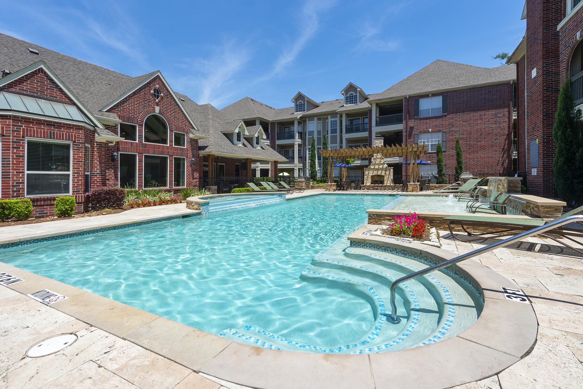 A spacious outdoor swimming pool surrounded by elegant brick apartment buildings. The pool features a shallow area with steps leading in, lounge chairs lining the sides, and vibrant landscaping with flowers. Bright blue skies add to the inviting atmosphere, making it a perfect spot for relaxation and leisure.