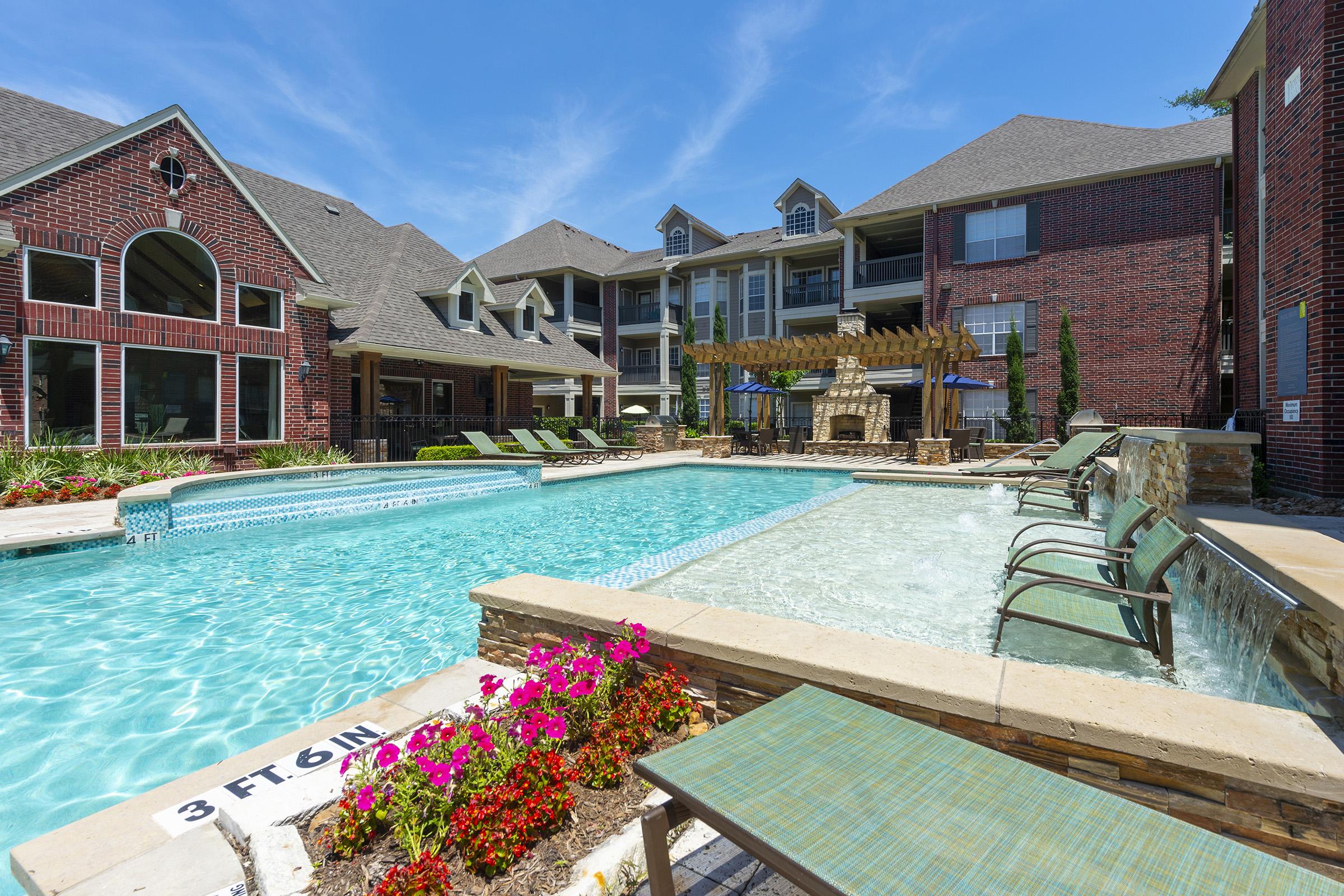 A serene swimming pool surrounded by lounge chairs and vibrant flowers, set within an upscale apartment complex. The area features brick buildings, well-maintained landscaping, and a cozy outdoor fireplace. Bright blue skies enhance the inviting atmosphere, perfect for relaxation and leisure.