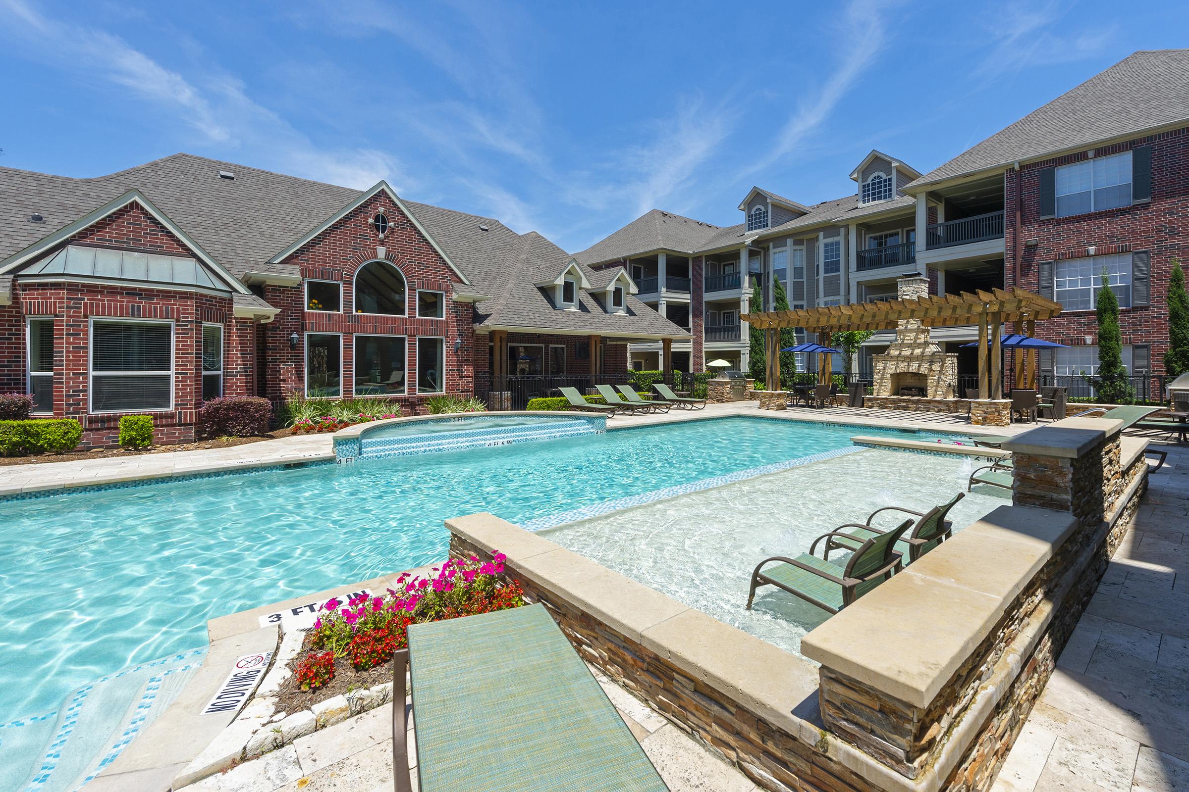 A bright, inviting pool area surrounded by brick buildings. The pool features lounge chairs, a shallow section with steps, and decorative landscaping with flowers. In the background, a multi-story residential building and a covered seating area with a fireplace are visible under a clear blue sky.