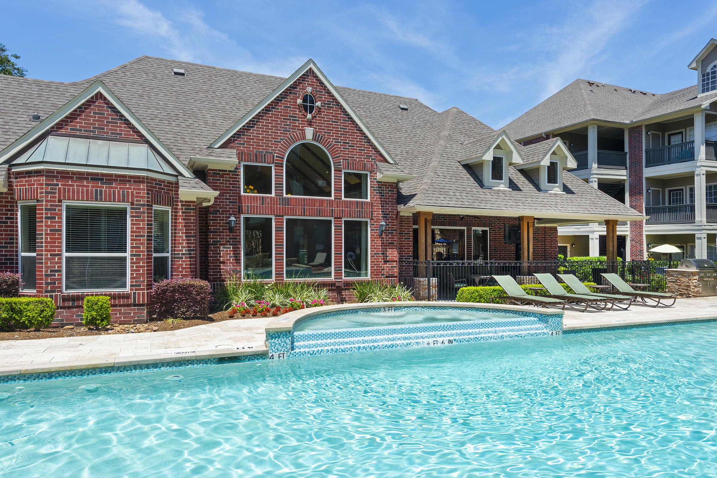 A beautiful outdoor scene featuring a large, sparkling swimming pool in the foreground, surrounded by lounge chairs. In the background, there is a stylish brick building with large windows and a covered patio, set against a clear blue sky. Lush greenery and colorful flowers enhance the inviting atmosphere.