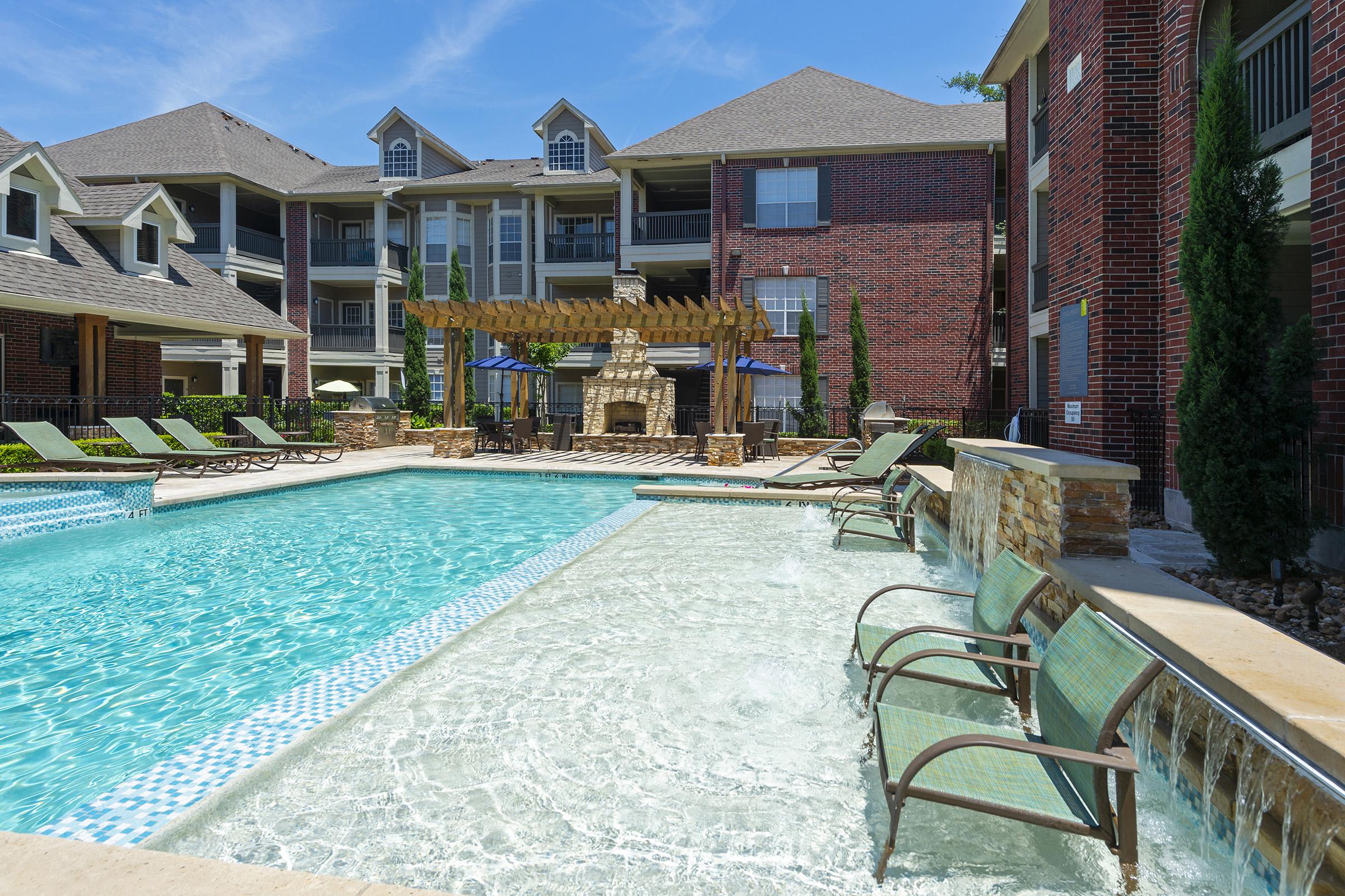 A view of a sparkling swimming pool with chaise lounges arranged around it. In the background, there are modern apartment buildings and a covered outdoor area featuring a stone fireplace and picnic tables. Lush greenery surrounds the pool area, creating a relaxing atmosphere on a sunny day.