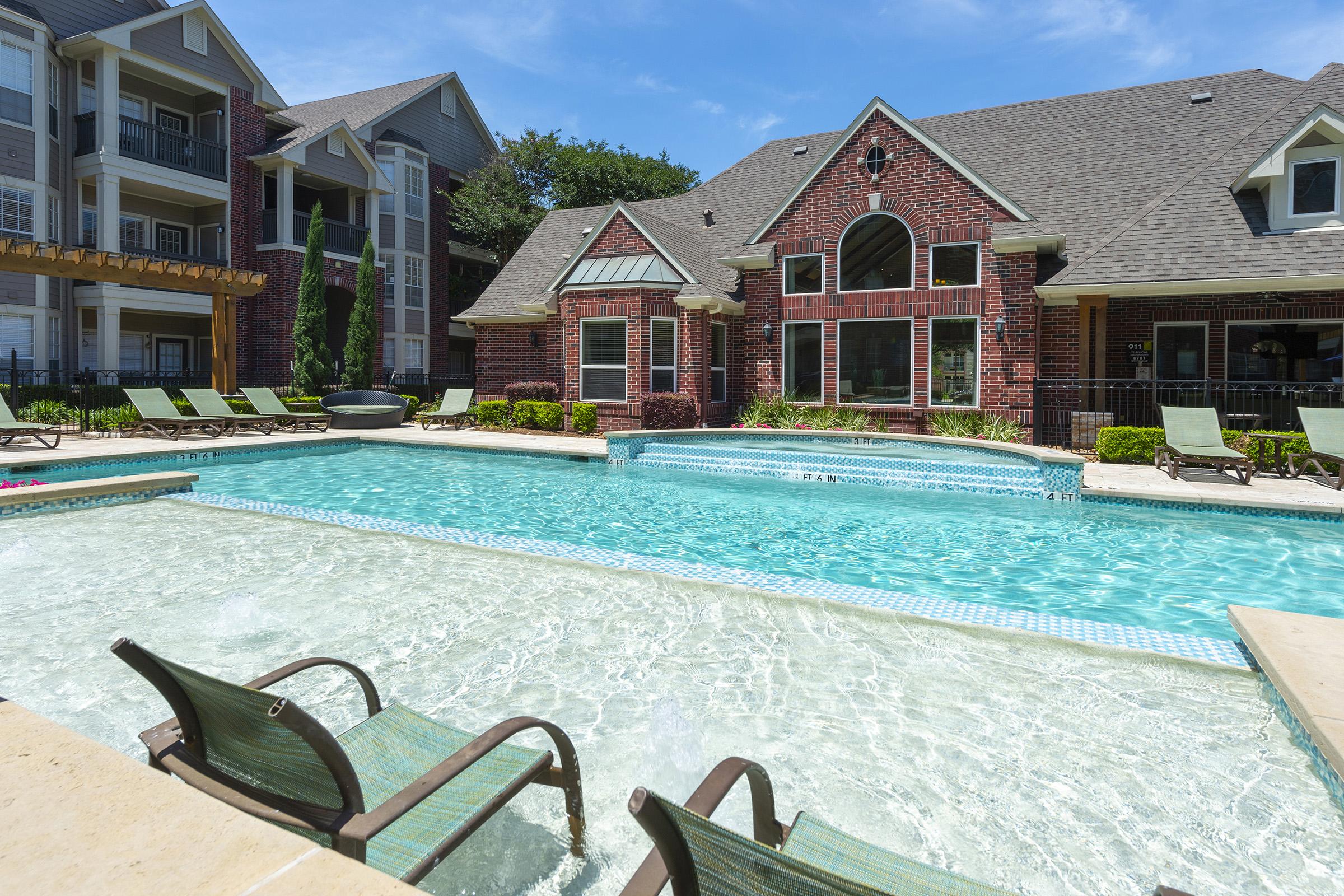 Bright blue swimming pool with clear water surrounded by lounge chairs. In the background, a brick apartment building features large windows and a well-maintained garden. The sky is clear with a few clouds, creating a relaxing outdoor atmosphere.