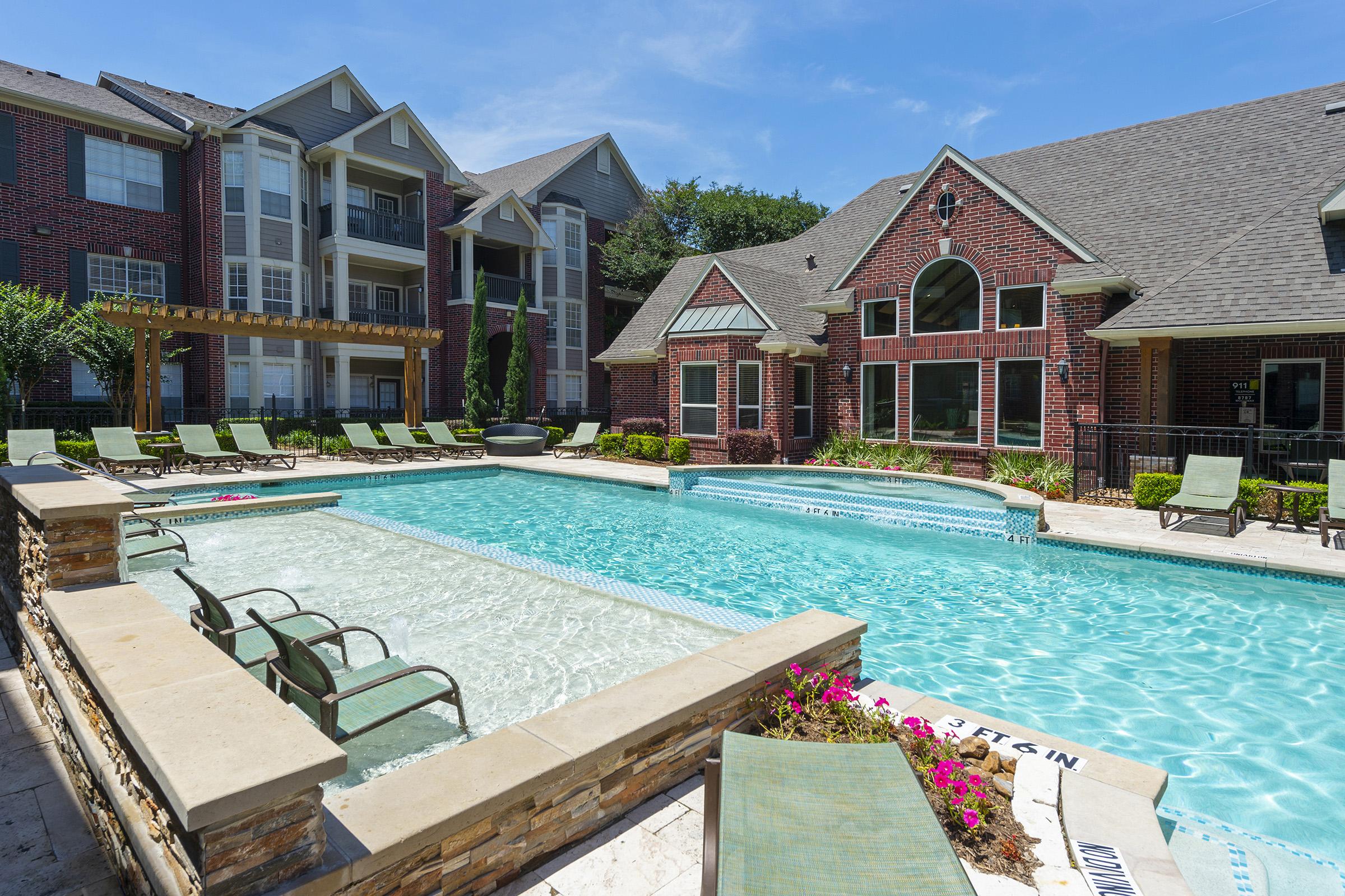 A sunny view of a swimming pool surrounded by lounge chairs and vibrant landscaping. The pool area features two sections, including a shallow area with a spa. Nearby, there are modern brick apartment buildings with large windows, creating a welcoming atmosphere for residents.